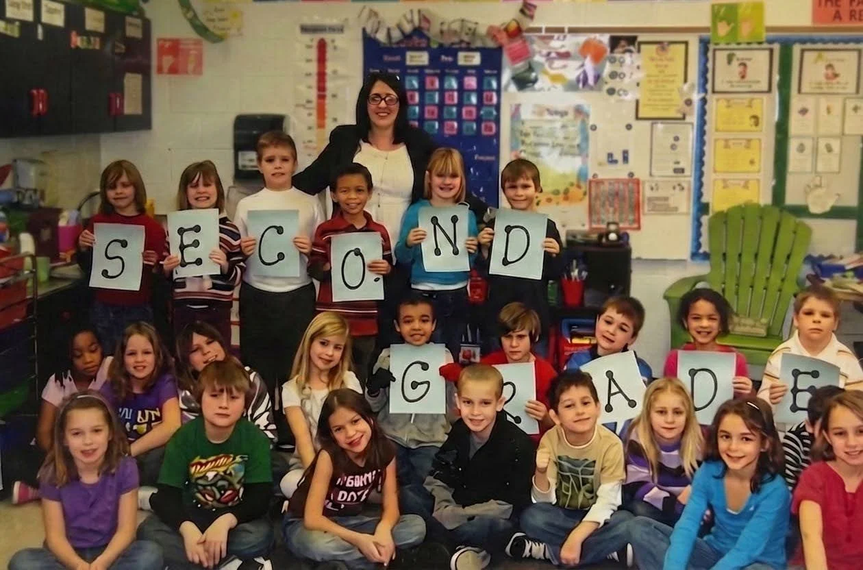 A classroom filled with young students and a teacher. The children are holding up signs that spell out "SECOND GRADE" and are smiling.
