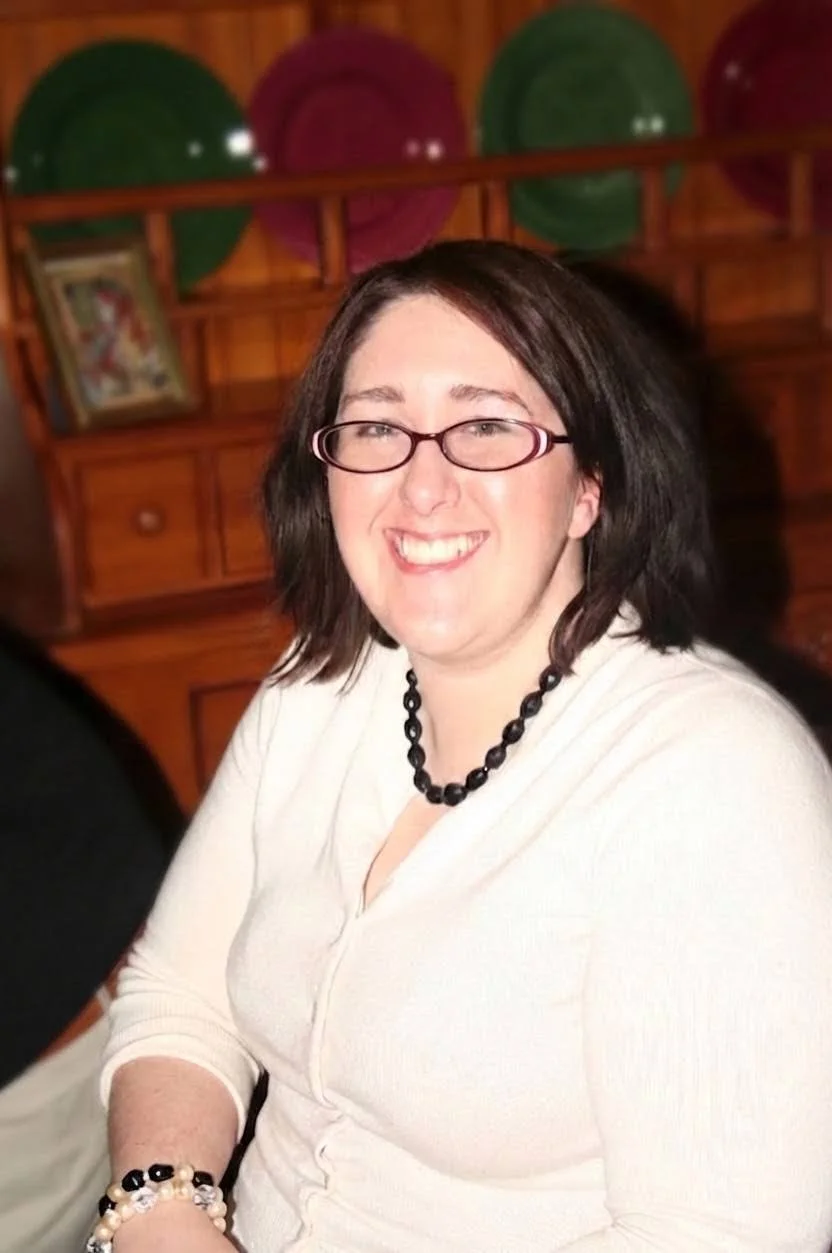 A woman with shoulder-length dark hair, glasses, and a white top, smiling, sitting in a room with a wooden wall and shelves behind her, decorated with green and pink balloons and a framed picture.
