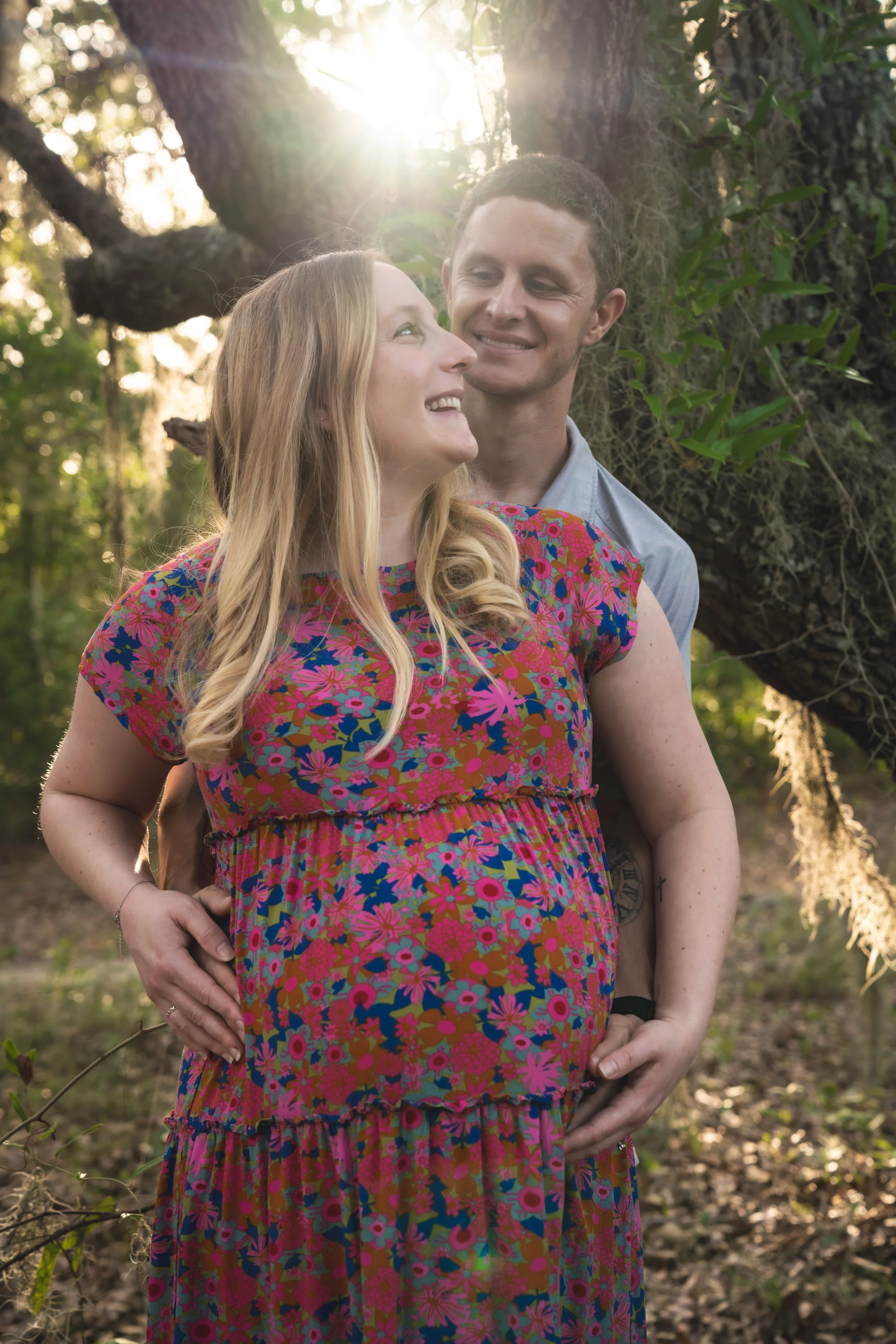 A pregnant woman wearing a colorful floral dress standing with a smiling man outdoors near a large tree with sunlight filtering through the leaves.