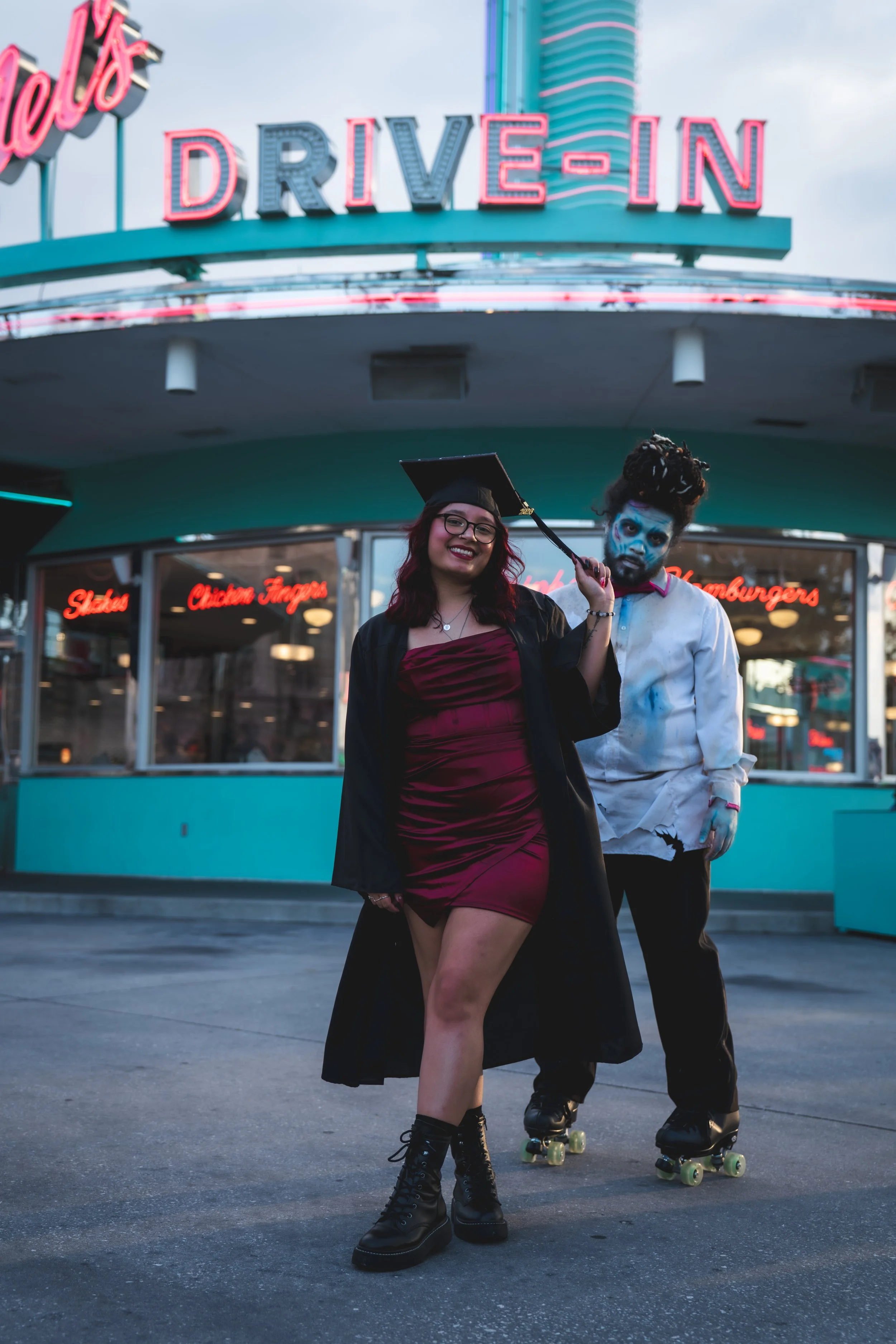 Two young adults, one woman in a maroon dress and black boots, and one man in a white shirt with makeup and dreadlocks, roller skating in front of a neon-lit drive-in restaurant at dusk.