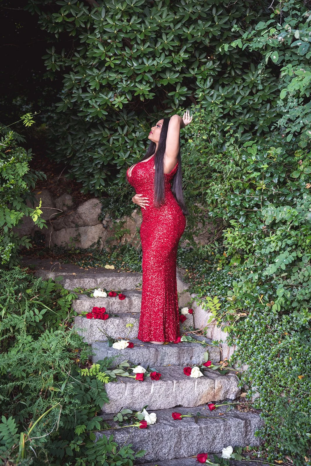 A woman in a red sequined gown standing on stone steps surrounded by greenery, with red and white roses scattered on the steps.