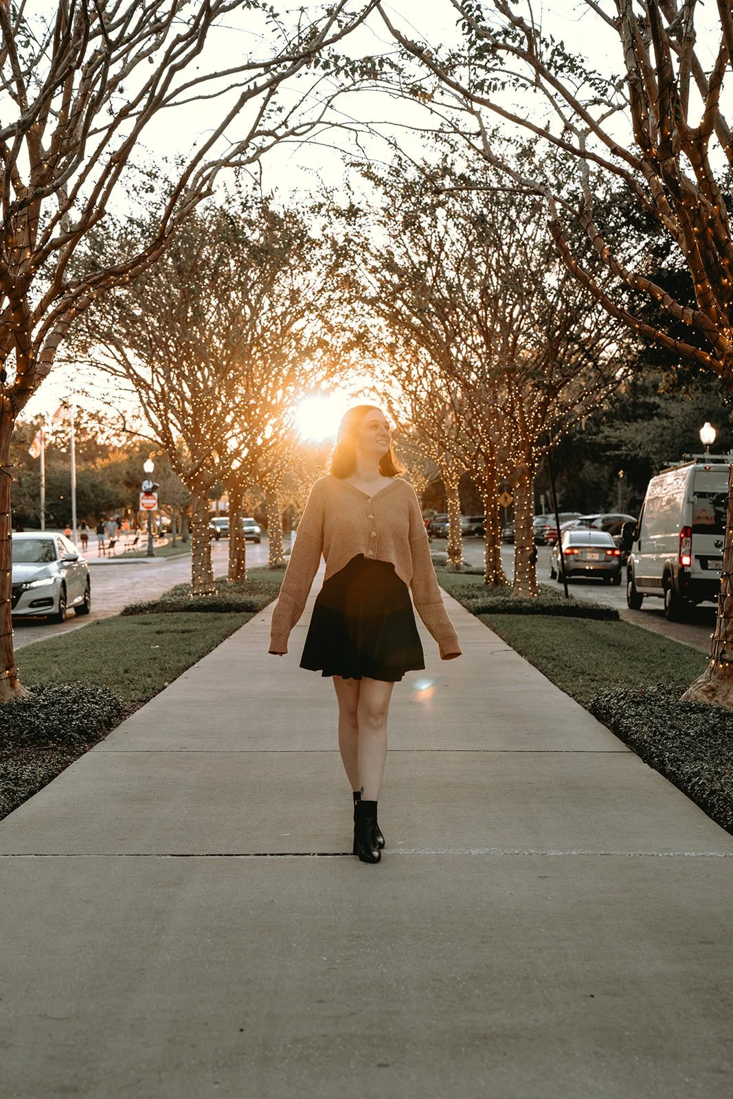 A woman walking on a sidewalk lined with trees decorated with string lights at sunset, with cars parked along the street.