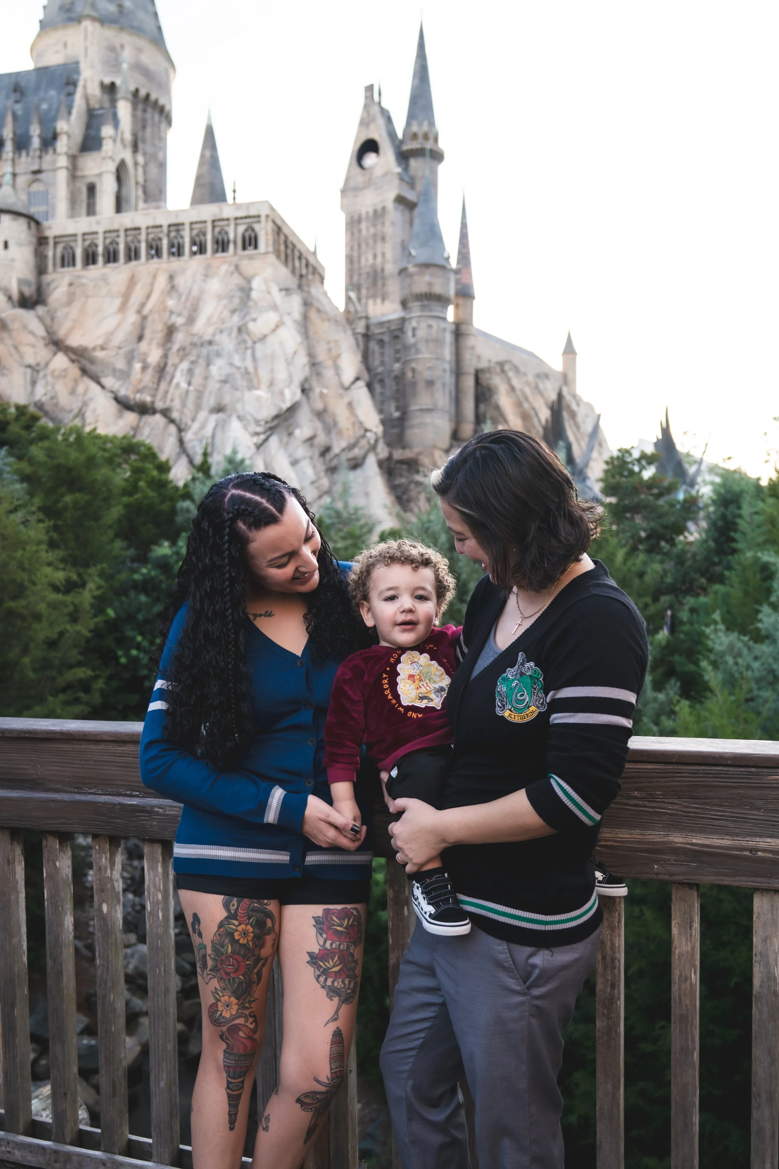 Two women and a young boy stand on a wooden viewing platform in front of the Harry Potter castle at Universal Studios, Orlando. The women are smiling and looking at the boy, who is sitting on one woman's lap, smiling. The castle is visible in the bac
