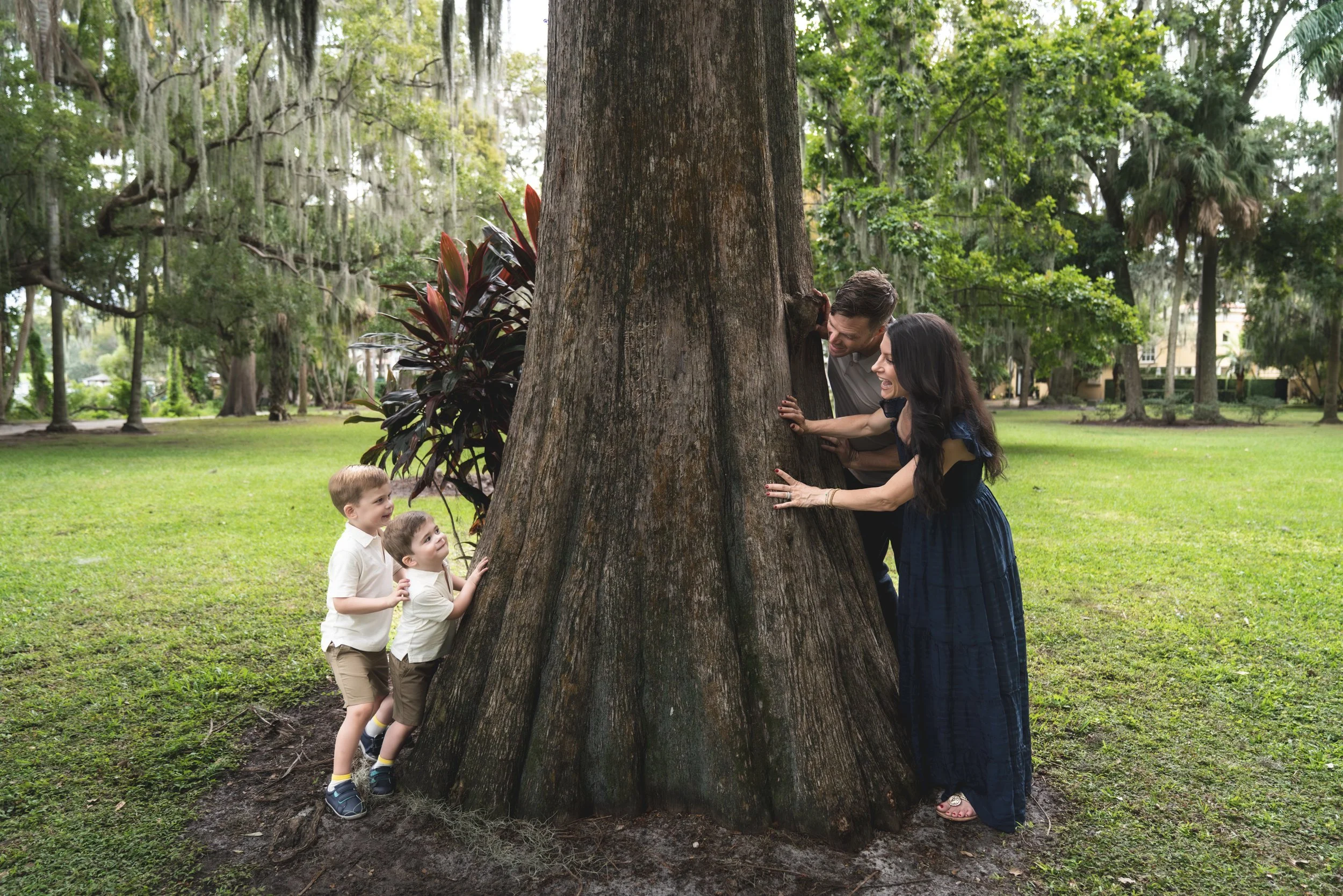 Family of five, including two children, hugging a large tree in a park with green grass and trees in the background.