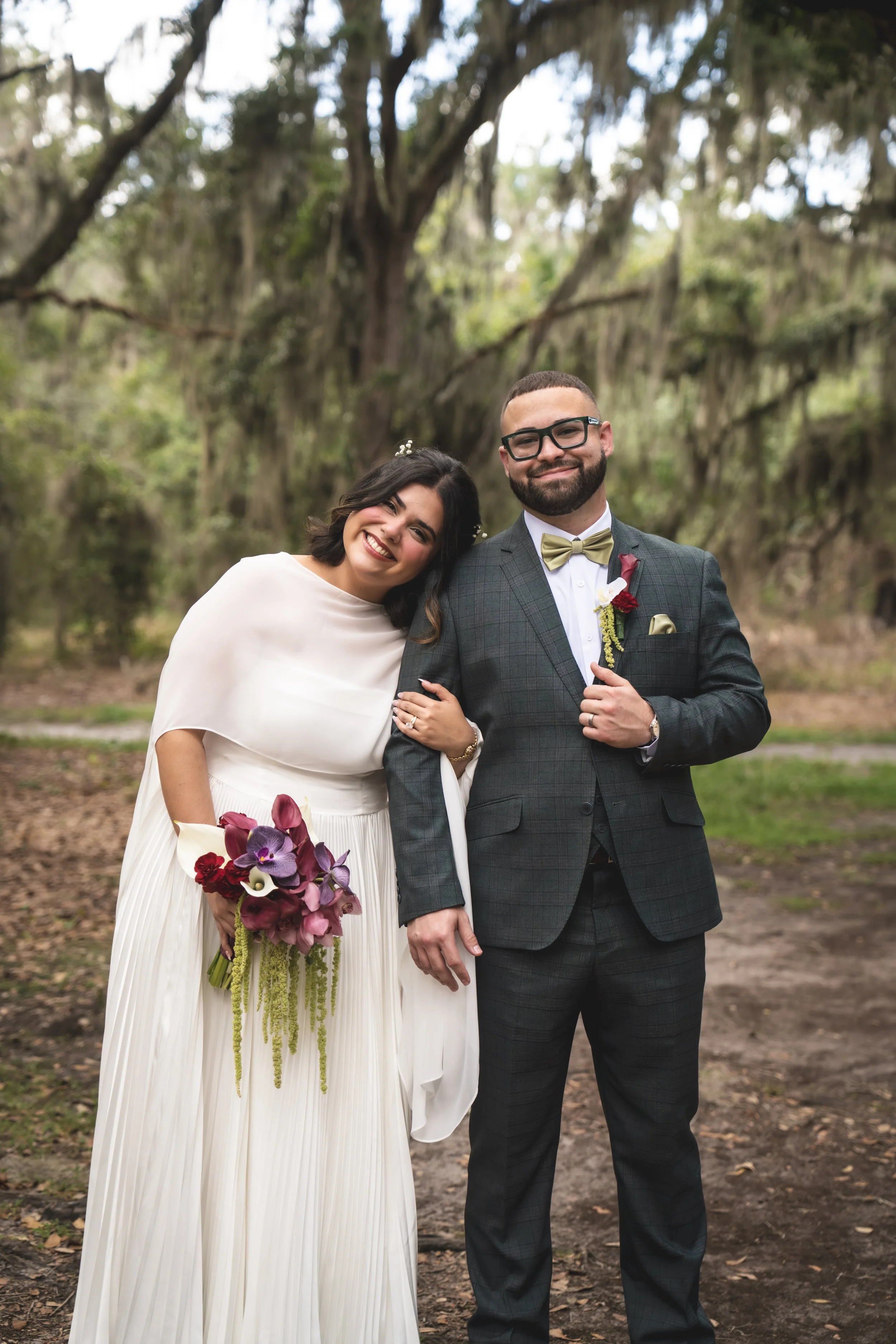 A happy bride and groom standing outdoors in a park-like setting with trees and moss hanging from branches, smiling at the camera. The bride is wearing a white gown and holding a bouquet of purple, red, and white flowers. The groom is wearing a gray suit with a bow tie and glasses.
