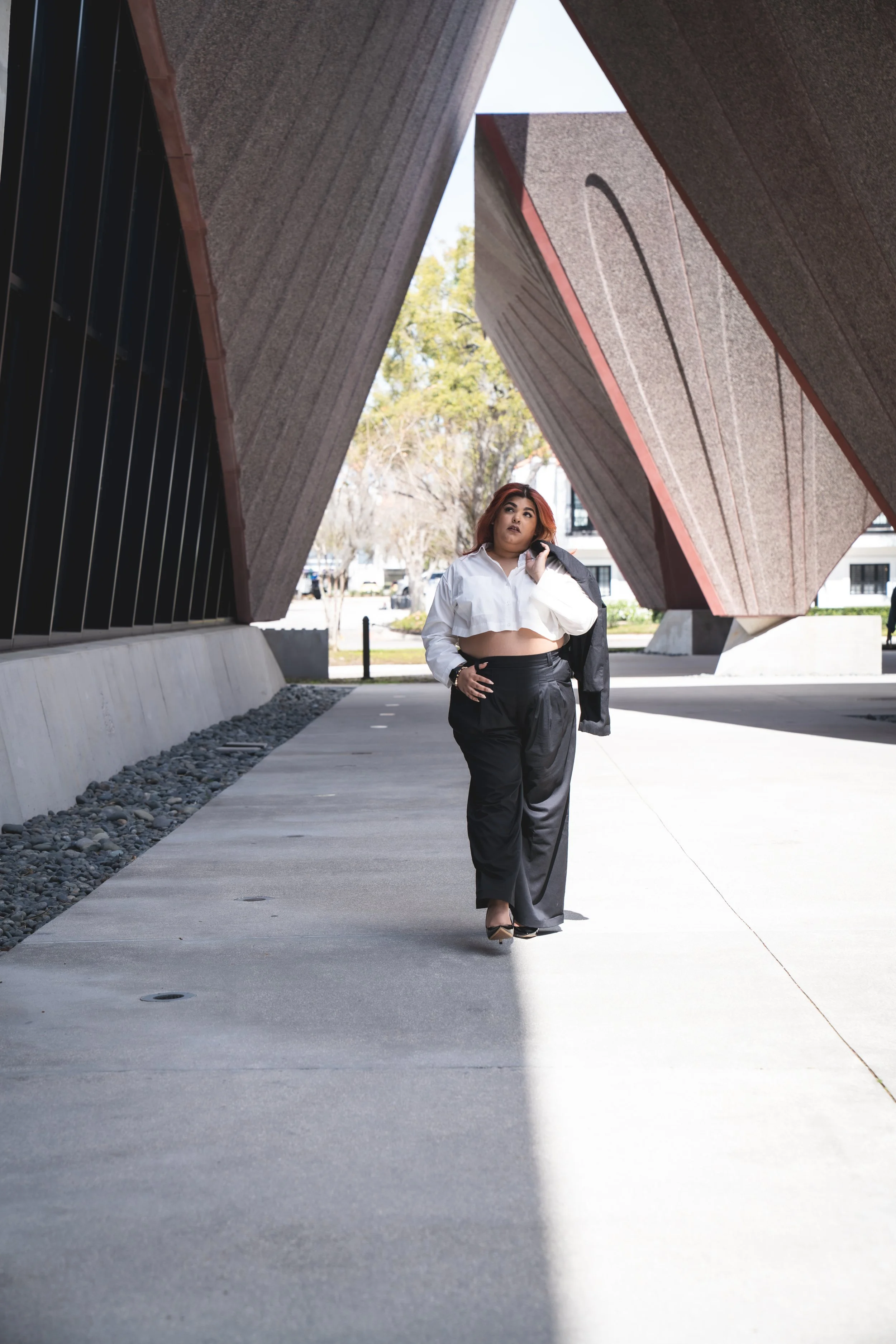 A woman with red hair, wearing a white crop top and black pants, walking on a sidewalk under modern, angular buildings.