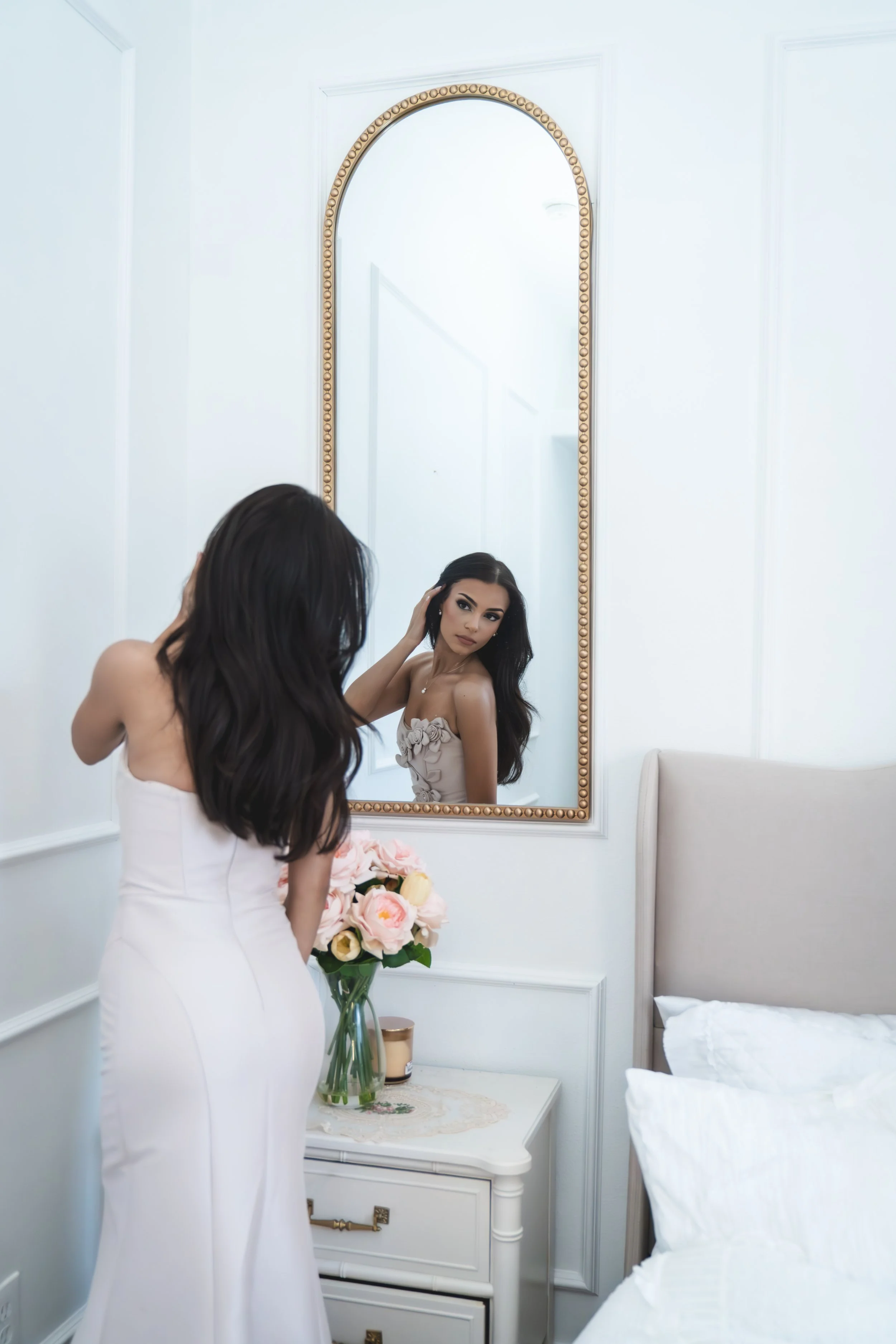 A woman in a white dress holding a bouquet of pink and cream roses, looking into a mirror in a light-colored bedroom.
