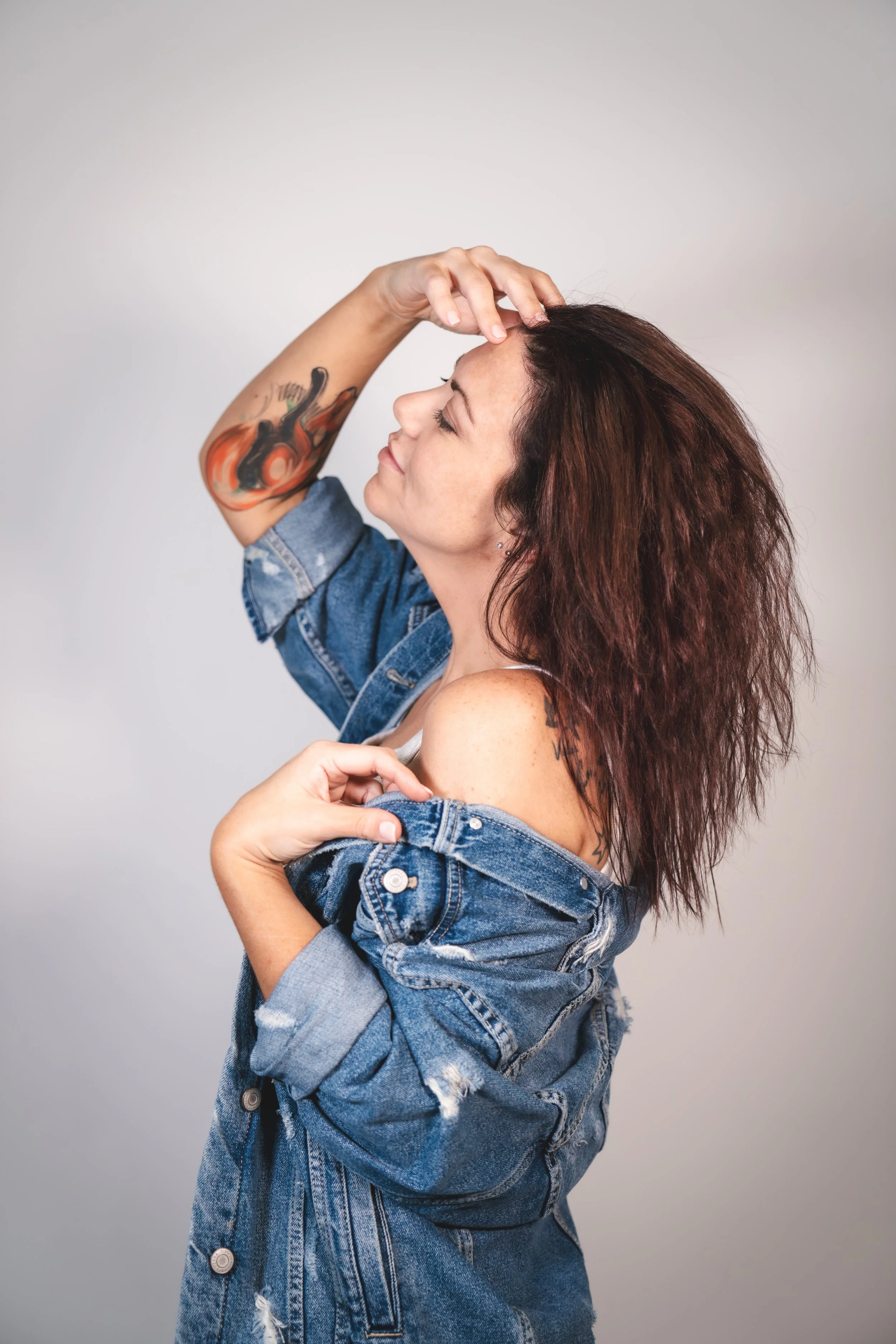 Woman with curly hair and tattoos on her arm, wearing a denim jacket, smiling with her eyes closed, holding her collar and touching her forehead, against a plain light gray background.