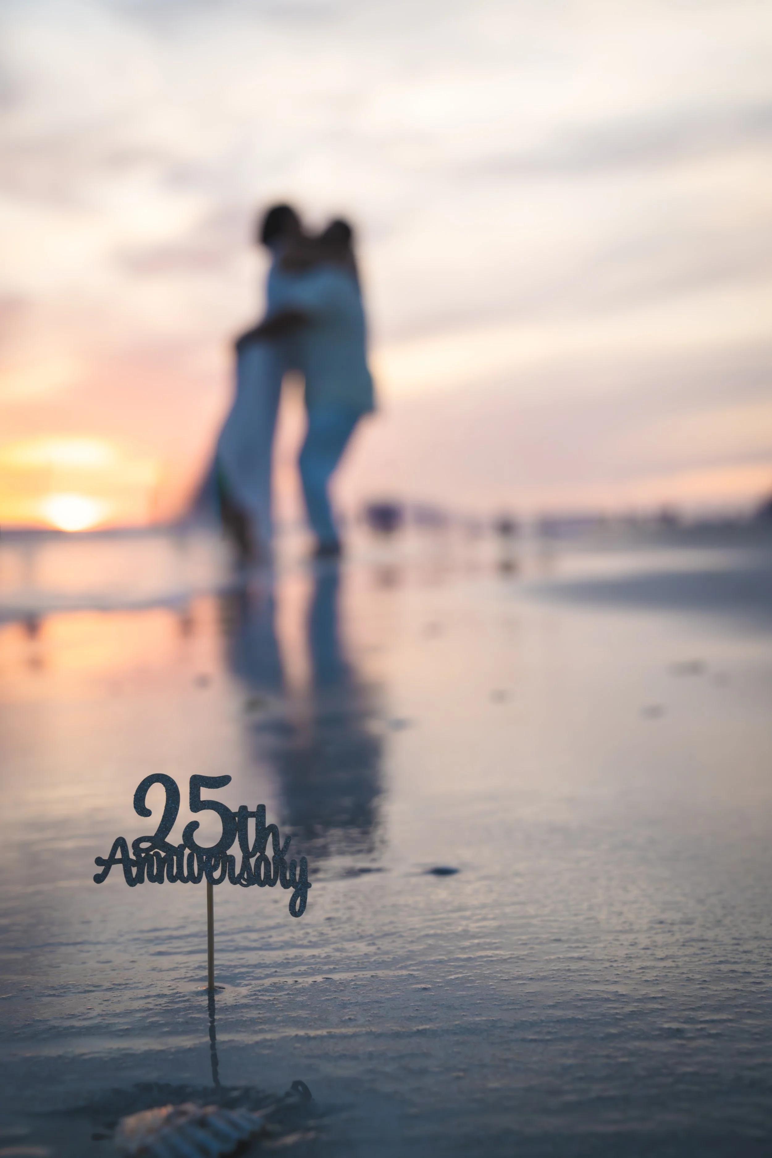 A couple walking on the beach during sunset, with a sign that says '25th Anniversary' in the foreground.