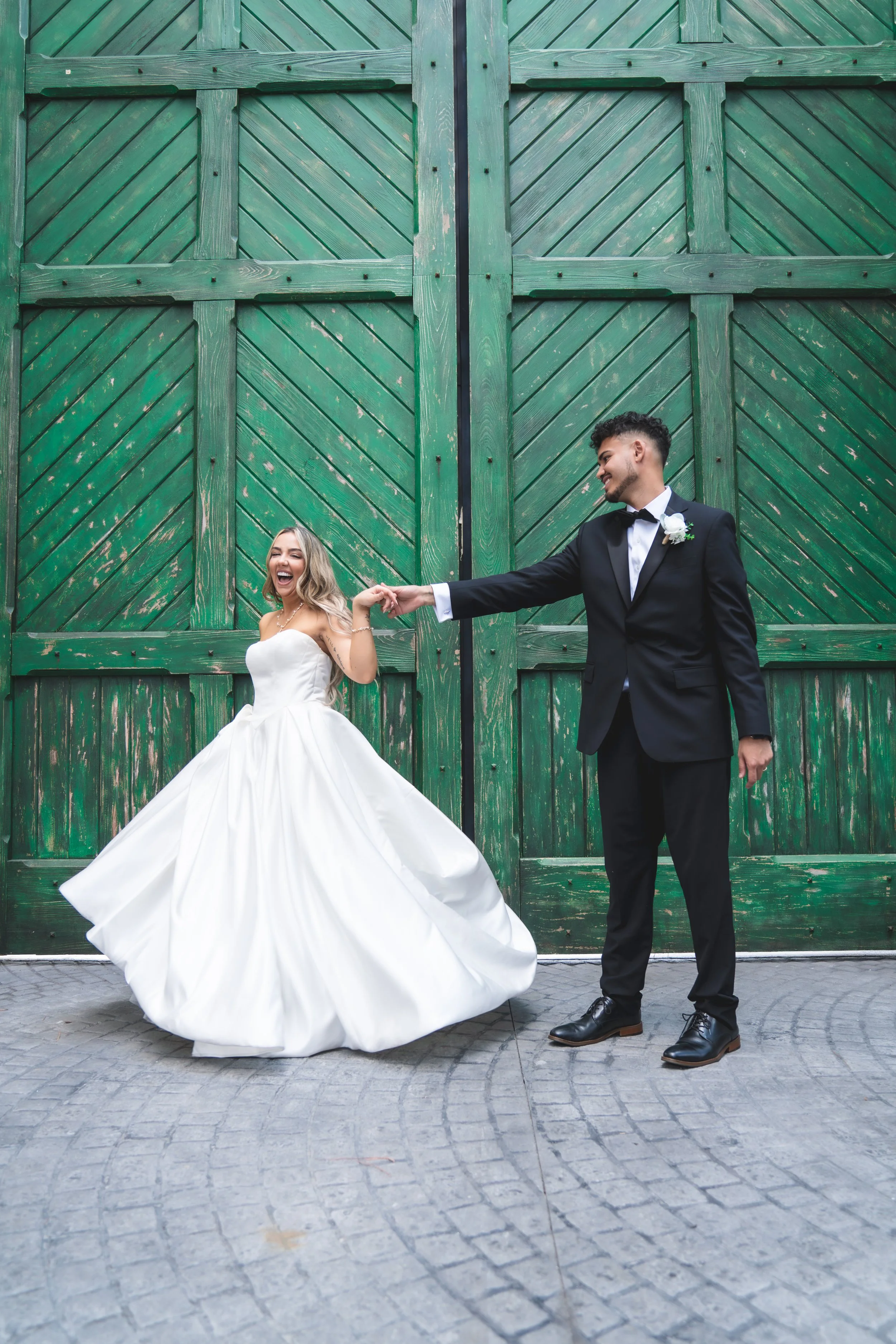 A bride and groom holding hands in front of a large green wooden barn door. The bride is wearing a strapless white wedding gown, and the groom is in a black tuxedo with a white shirt and black bow tie, smiling at each other.