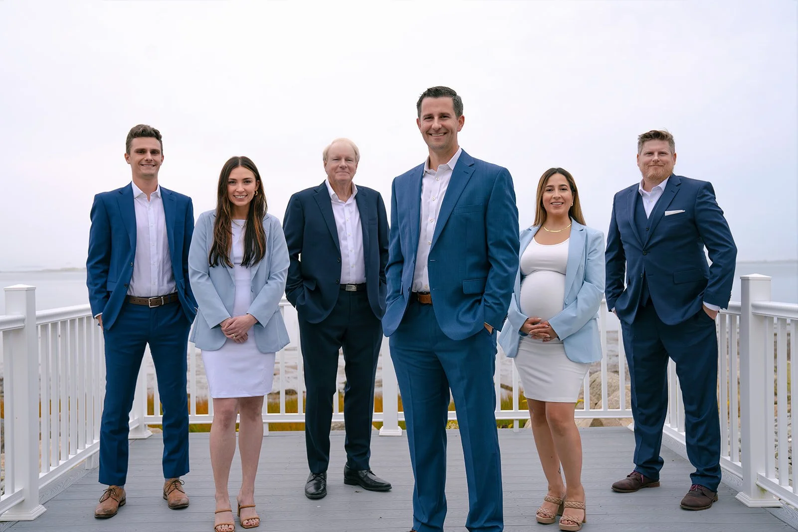 Group of six professionally dressed people standing outdoors on a deck by the water, smiling at the camera.