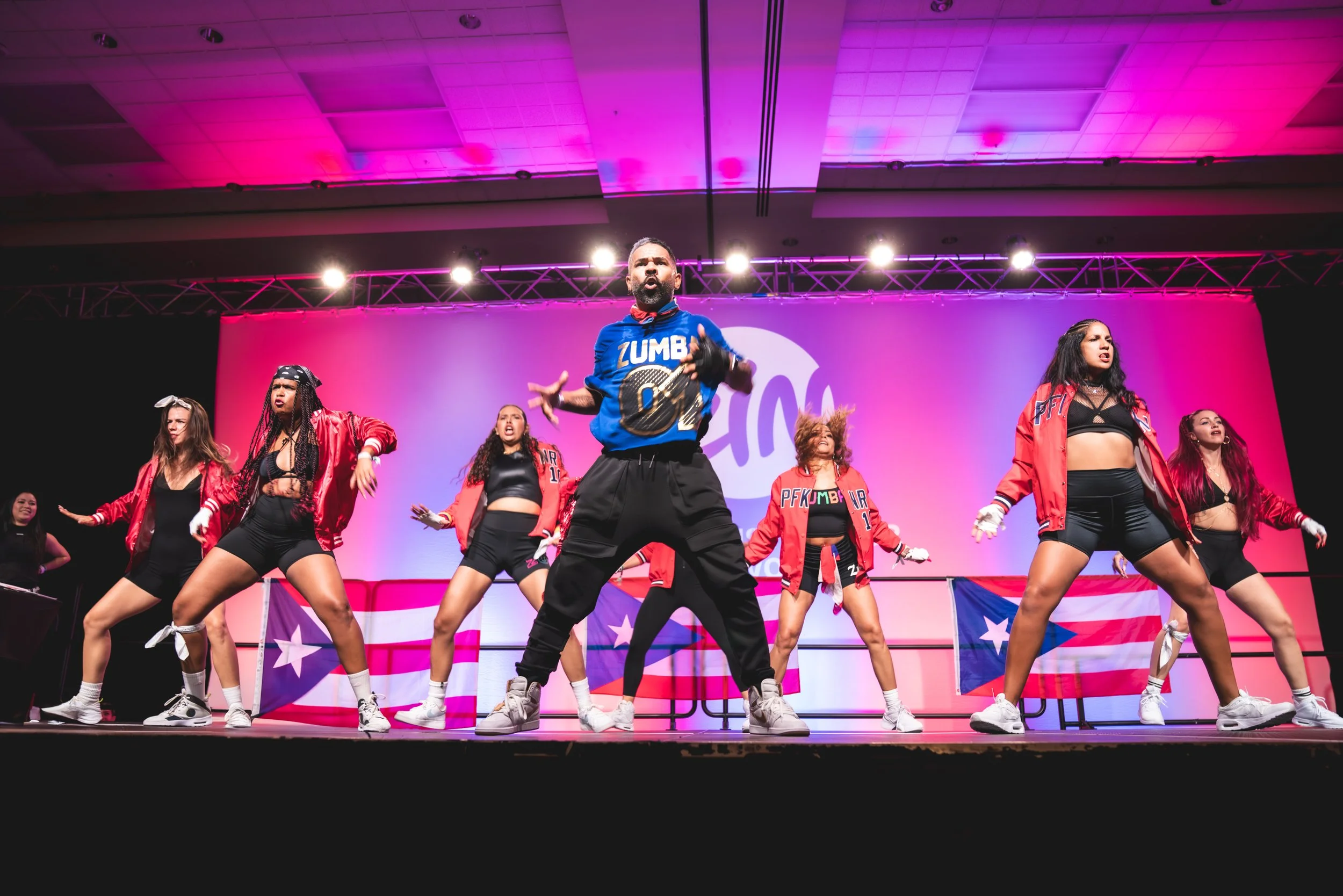 A group of dancers performing on stage at a Pink Nation event with purple and pink lighting. They are dressed in colorful outfits and are in choreographed poses.