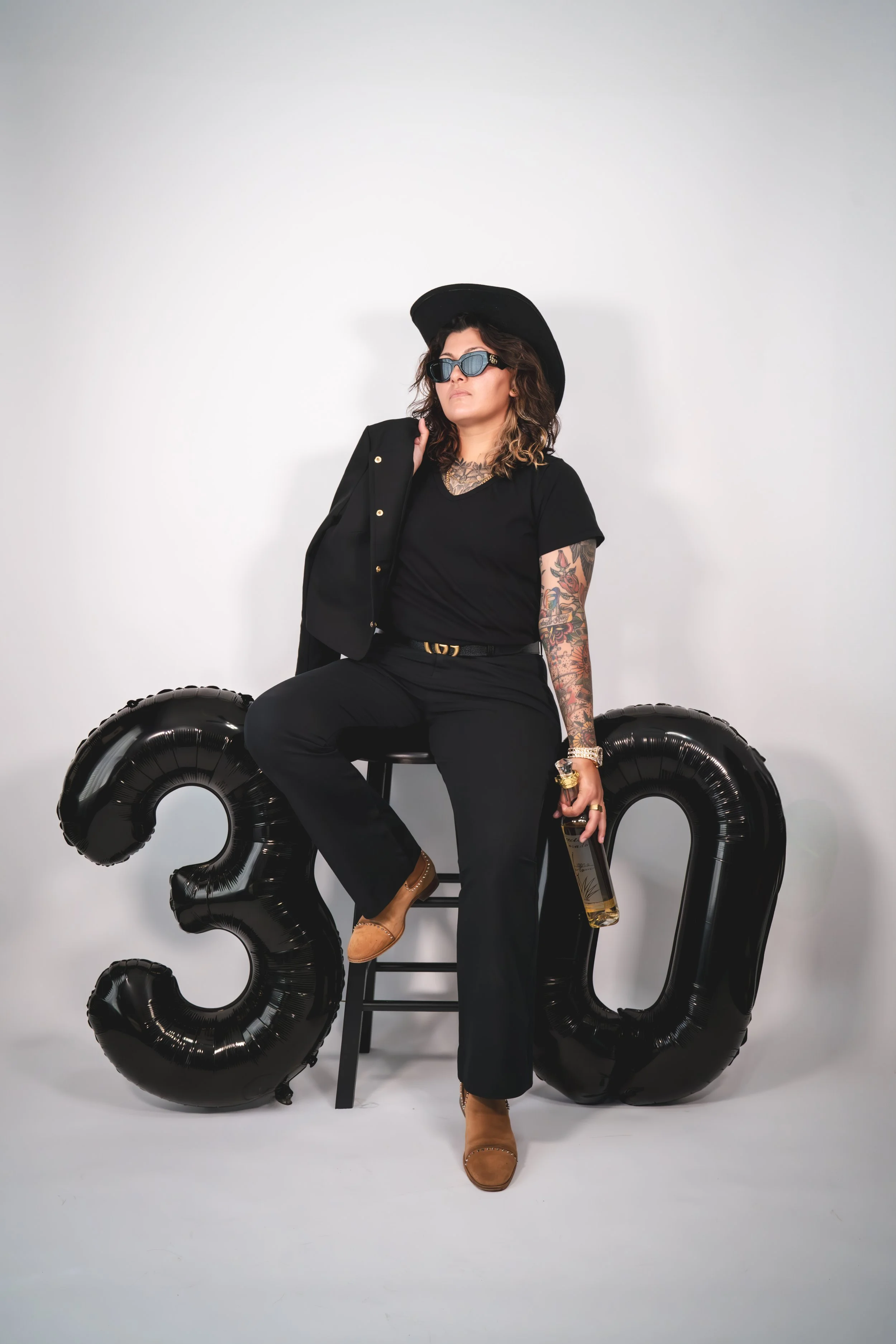 A woman with tattoos, wearing sunglasses, a black hat, black shirt, and black pants, sitting on a black ladder and holding a bottle of wine, with large black balloons shaped as the number 30 behind her against a plain white background.