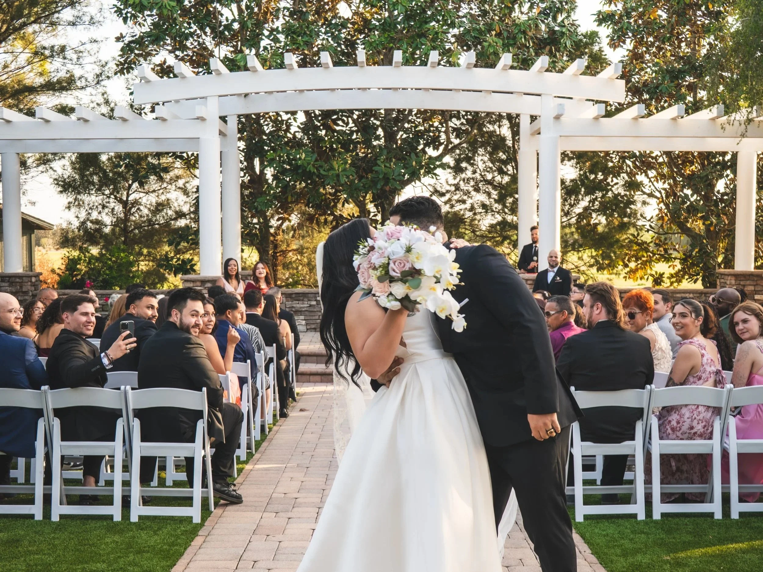A wedding ceremony outdoors with couple kissing and embracing while holding a bouquet of white and pink flowers, surrounded by seated guests and a white pergola in a garden setting.
