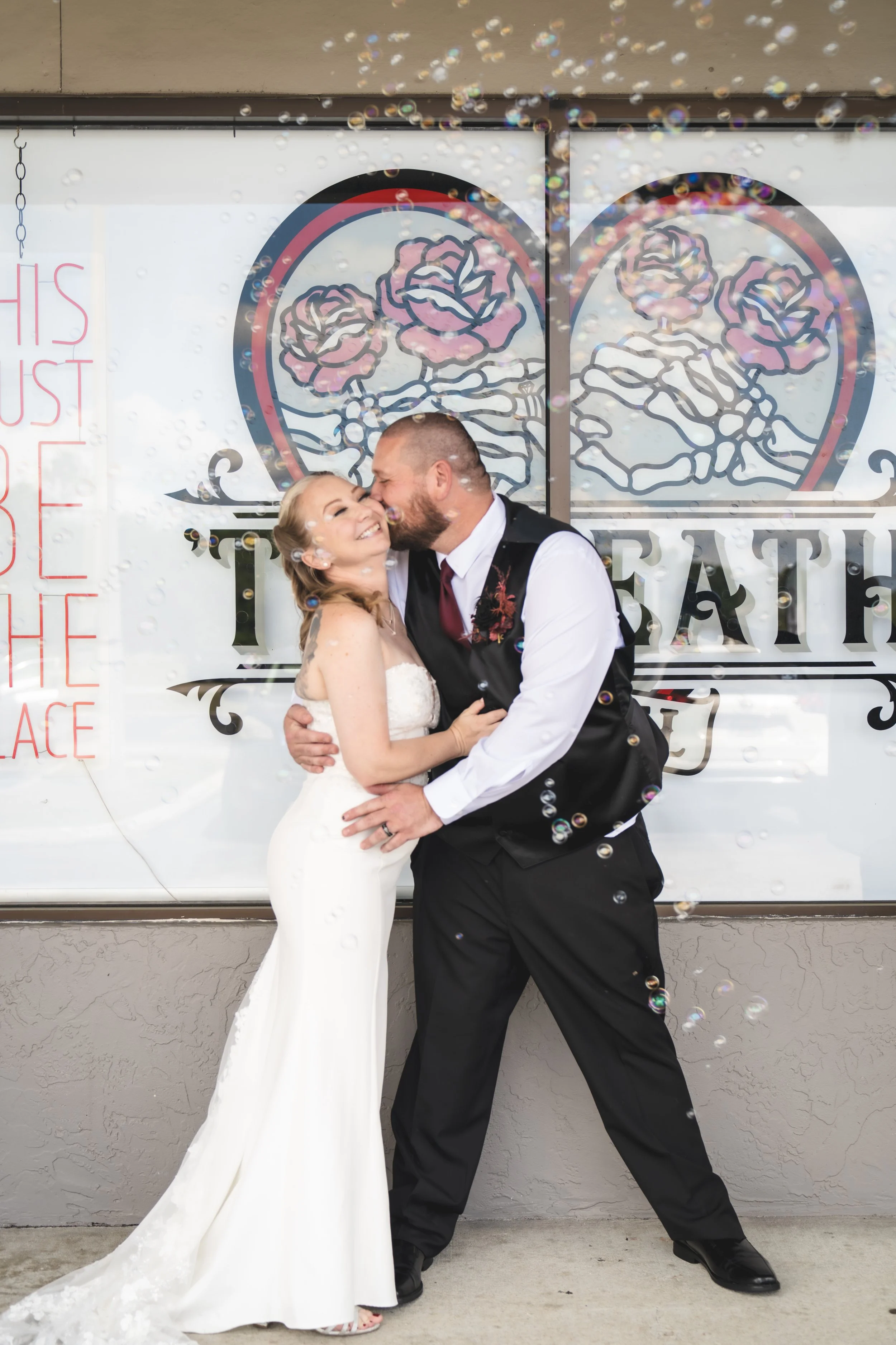 A newlywed couple, the bride in a white gown and the groom in a black vest and pants, sharing a kiss and embrace in front of a decorated backdrop with a heart and roses design, with bubbles floating around them.