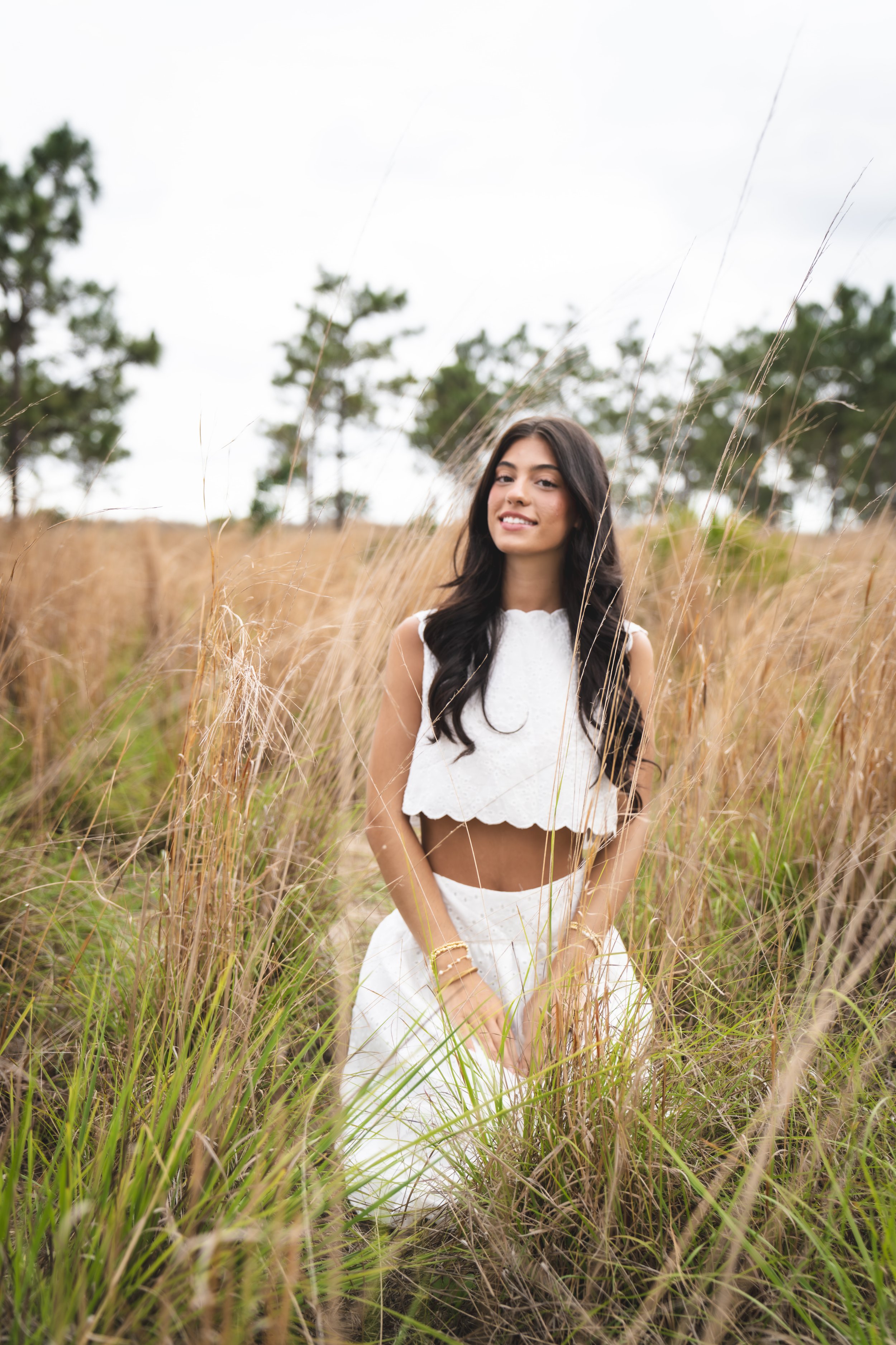 A young woman with long black hair and a white crop top standing in a field of tall, dry grass with a few trees in the background, under an overcast sky.