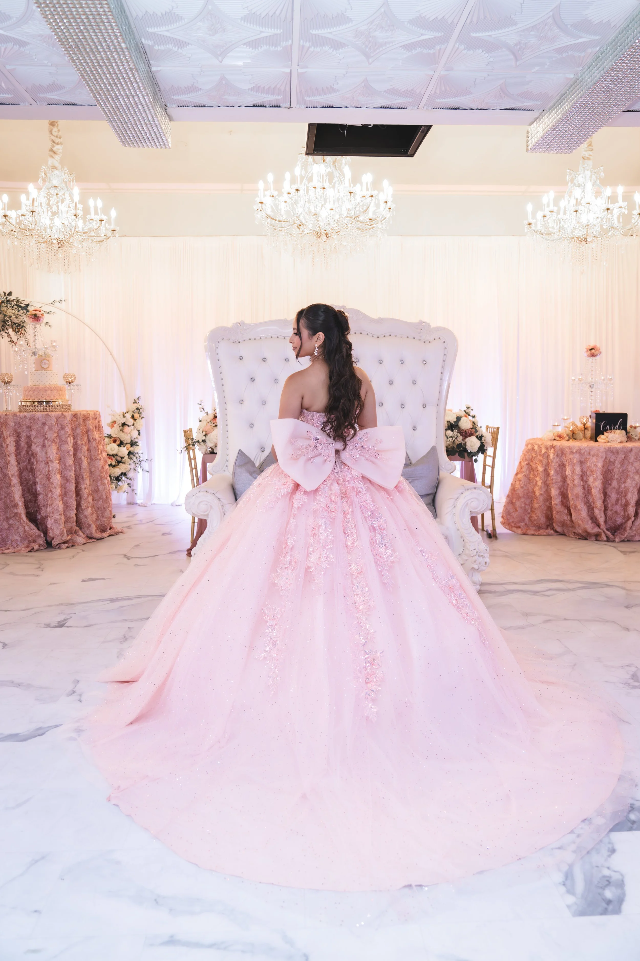 A woman in a pink prom dress sitting on a white ornate throne, surrounded by elegant chandeliers, floral arrangements, and decorated tables in a luxurious event hall.