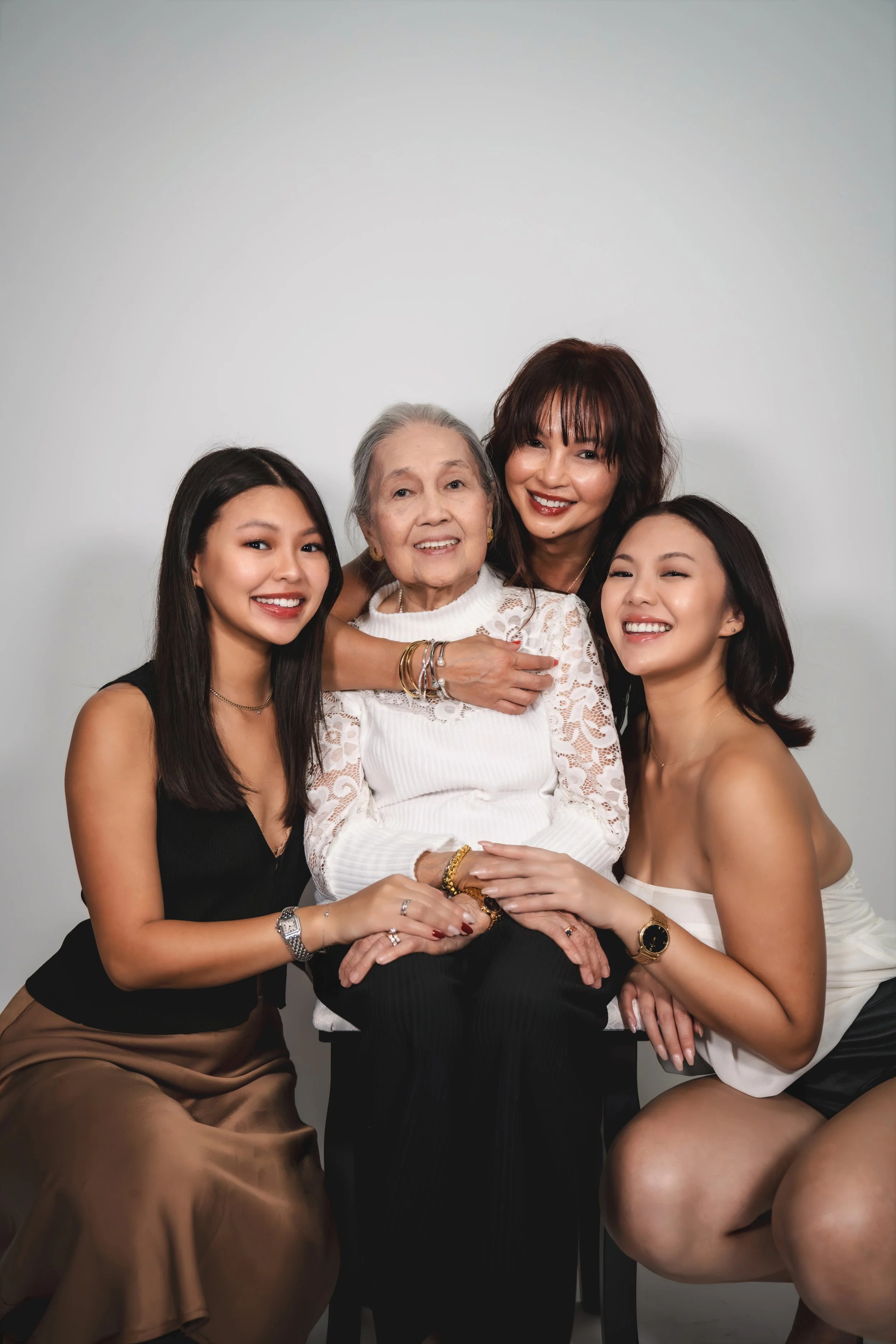 Four women of different ages and ethnicities posing together against a plain light gray background, smiling and embracing each other.