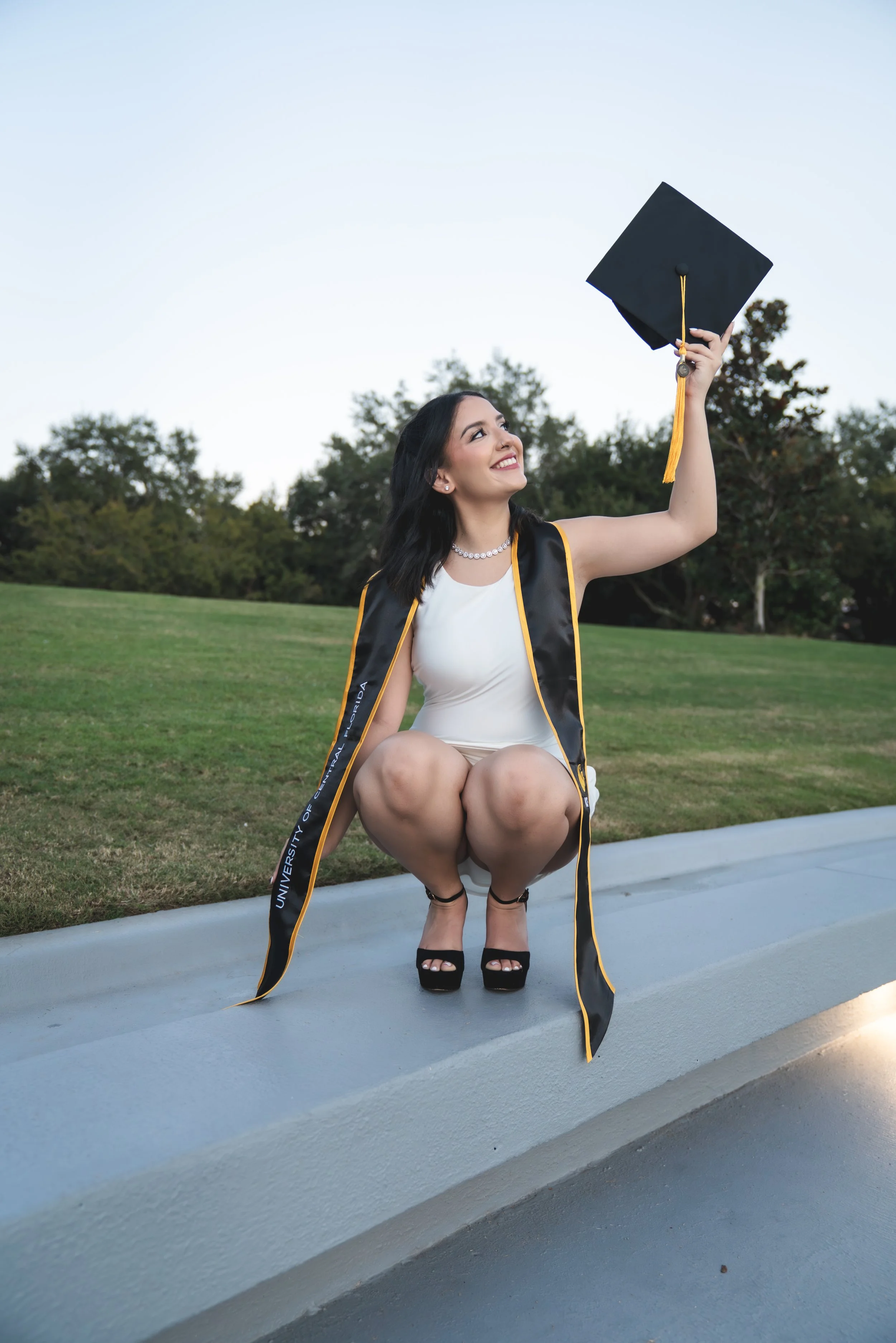 A young woman sitting on a low wall outdoors, wearing a graduation cap and gown, holding her cap in the air with a smile, with trees in the background on a clear day.