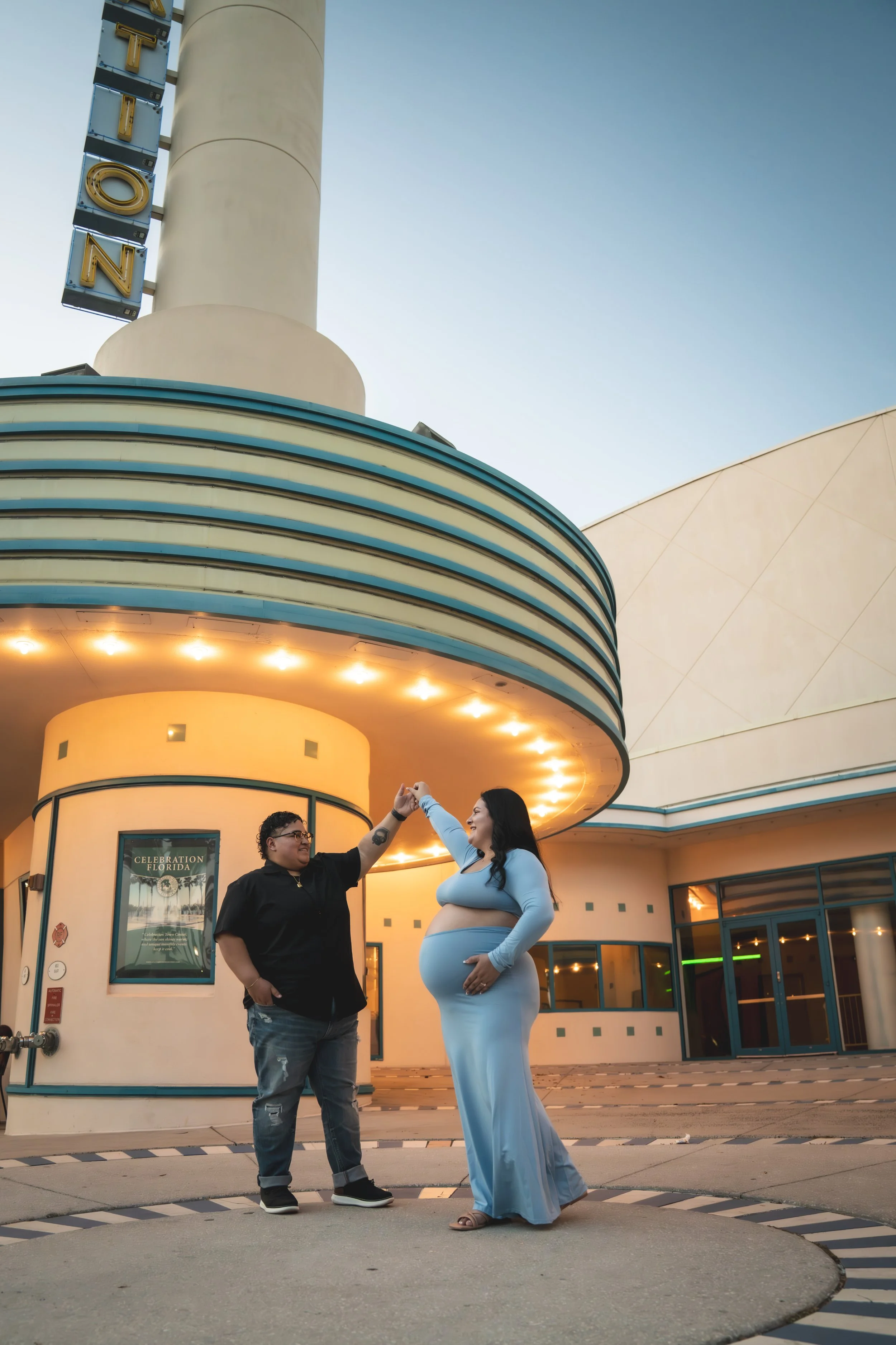 Two people dancing in front of a movie theater, with one person appearing pregnant, the other person wearing glasses, both smiling and holding hands, under a cloudy sky.
