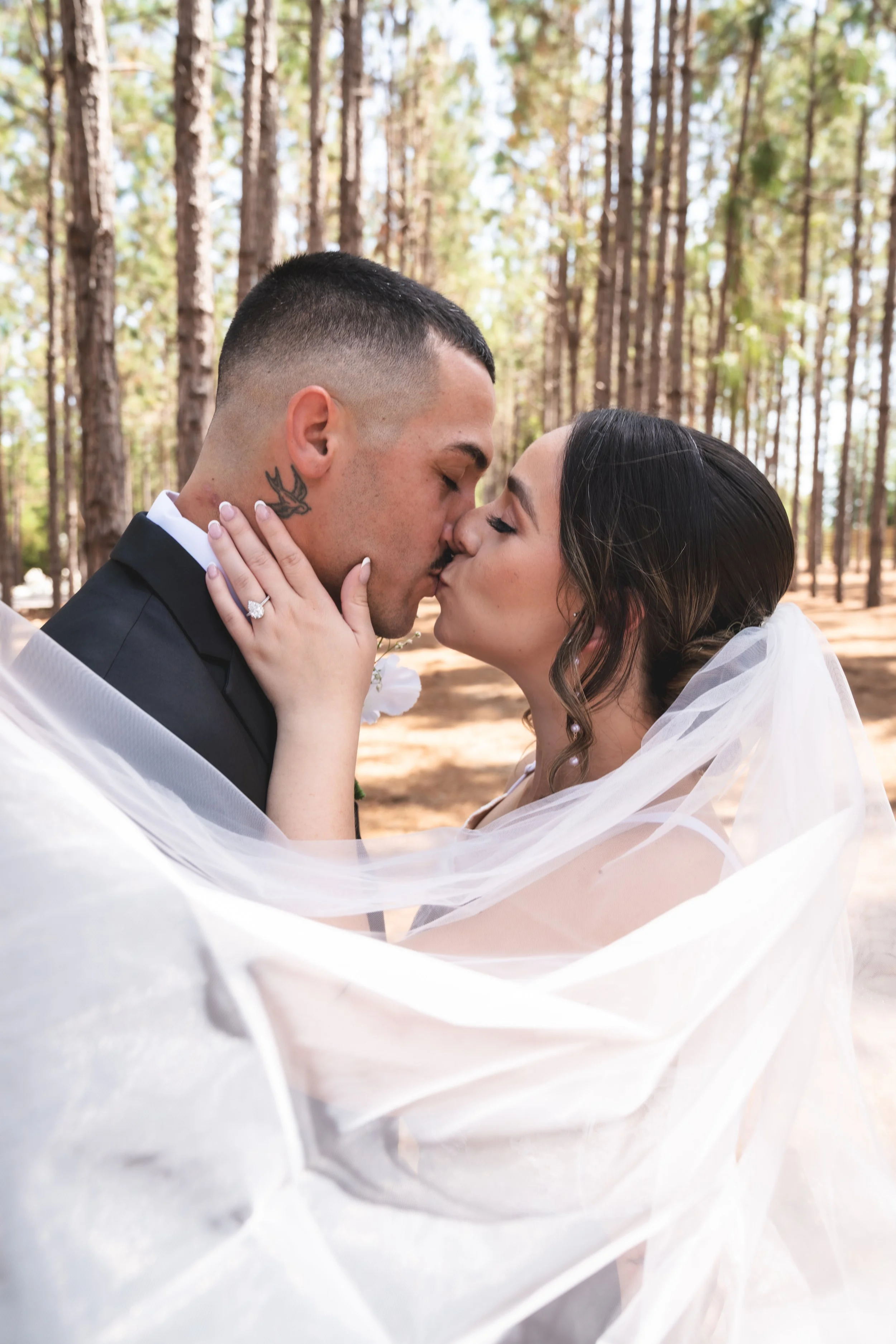 A couple kissing outdoors in a forest with trees in the background, dressed in wedding attire.