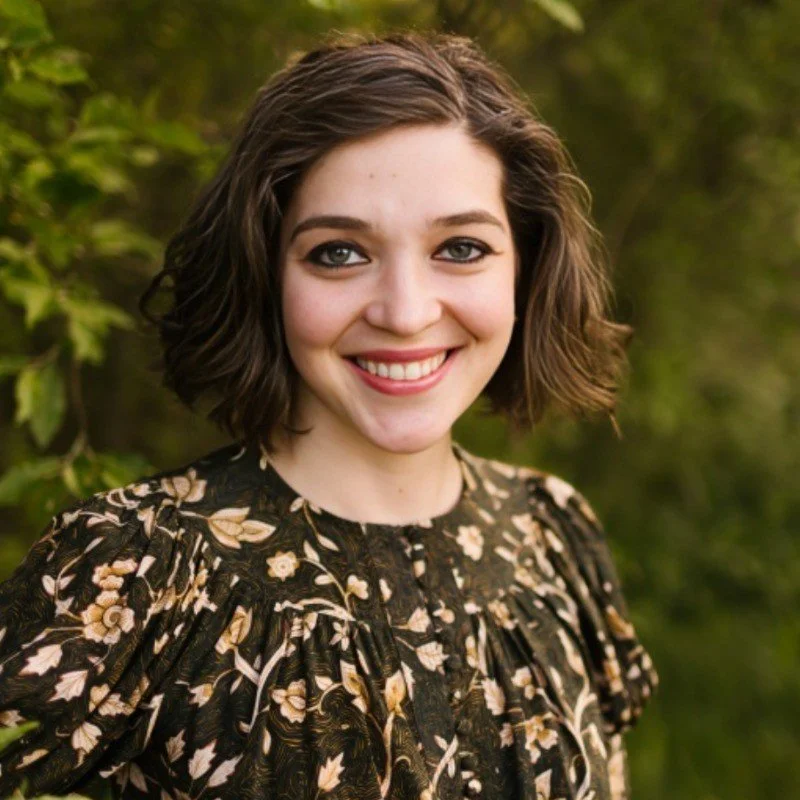 An image of Katie Bauer, a brunette woman facing toward the camera and smiling in front of a green tree