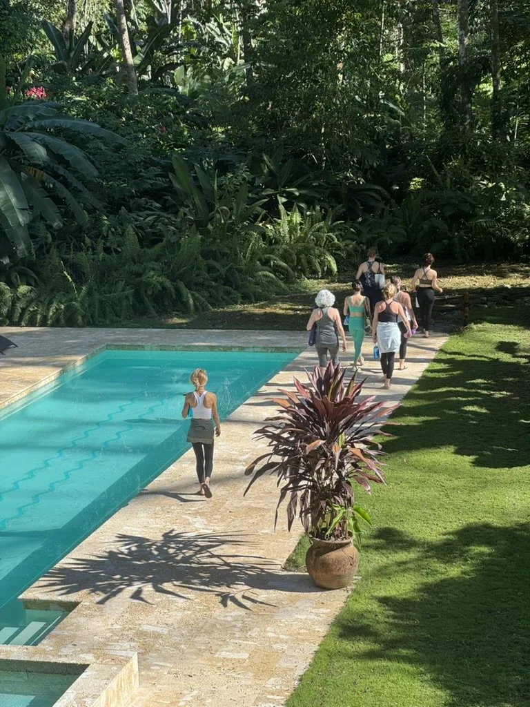 A woman walking beside a swimming pool with a group of women walking toward the forest or garden area in the background, surrounded by lush green plants and trees.