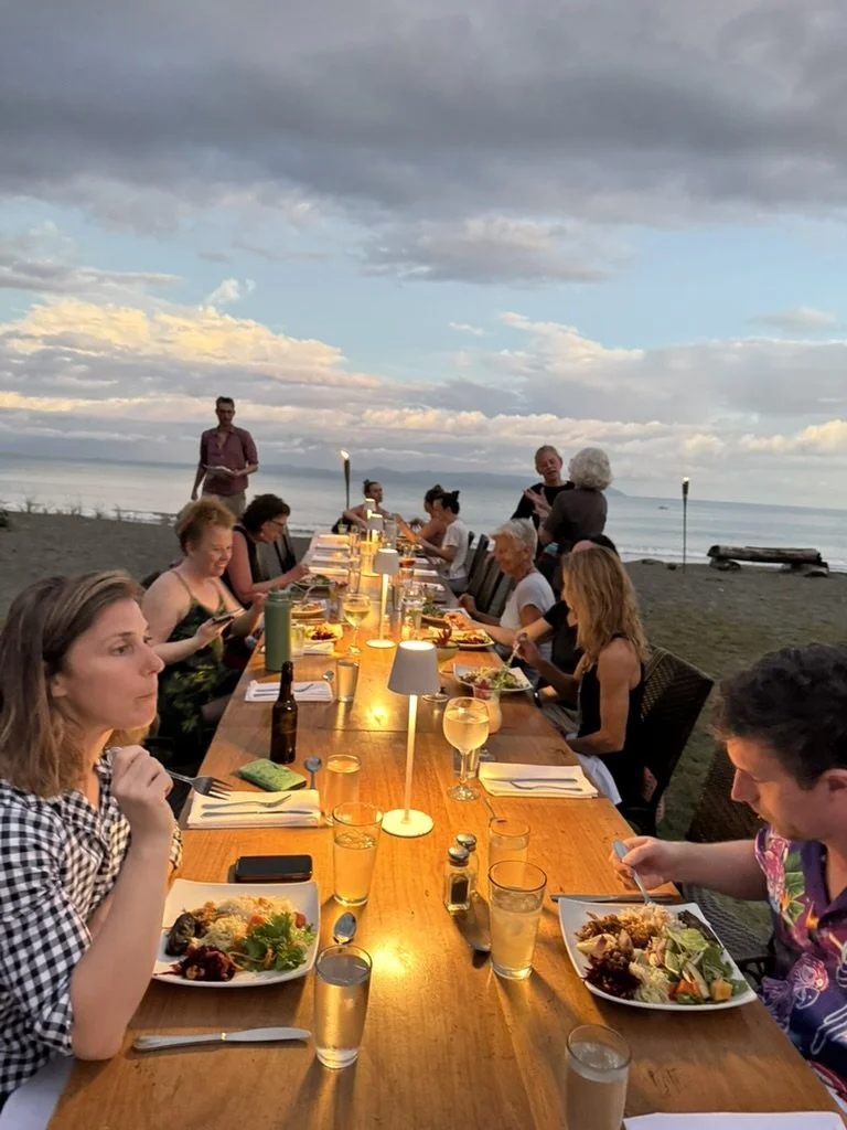 People dining at a long outdoor table on a beach during sunset, with the ocean and sky in the background, and lit lamps along the table.
