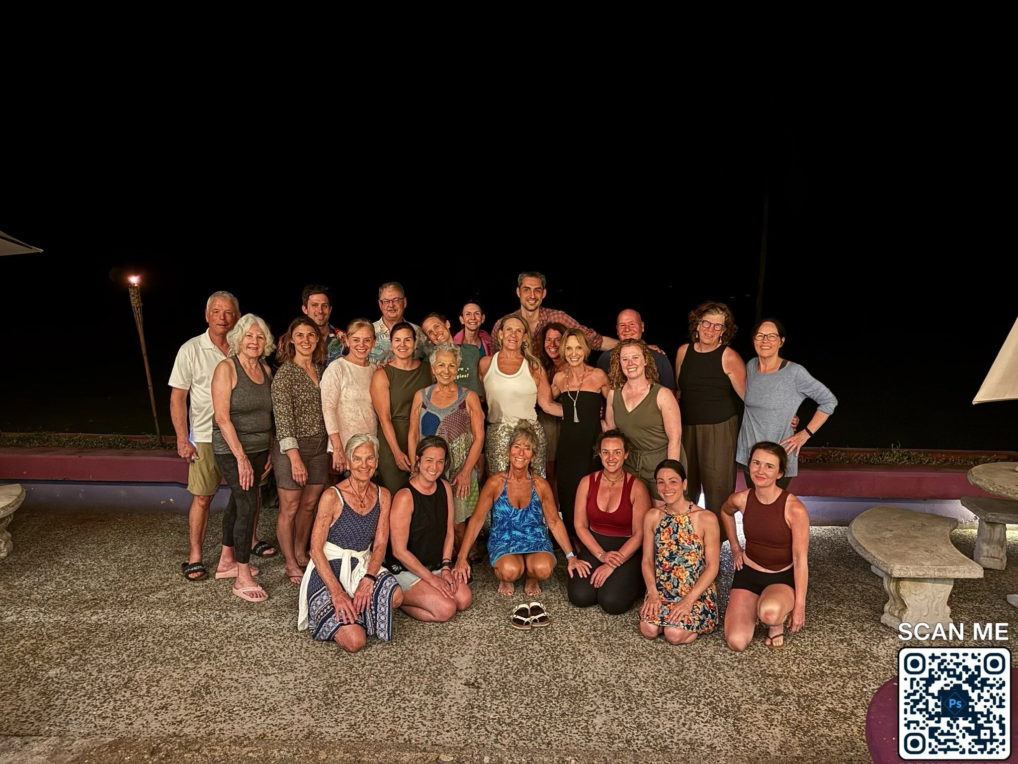 A group of 23 people gathered outdoors at night, posing for a photo with a dark sky background. The group is smiling and standing or sitting on a textured ground, with two stone benches and umbrellas visible.