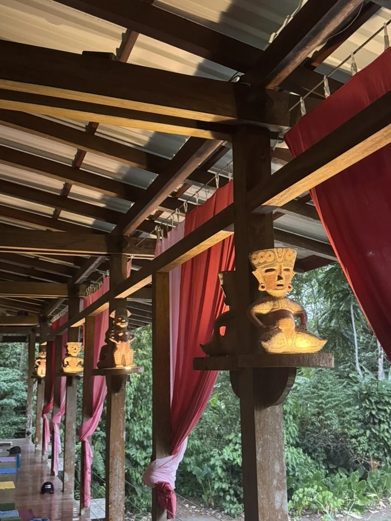 Wooden beams supporting a structure with red curtains hanging between the beams. Traditional carved wooden figures, resembling indigenous or tribal art, are mounted on the beams. The background shows lush green foliage outside.