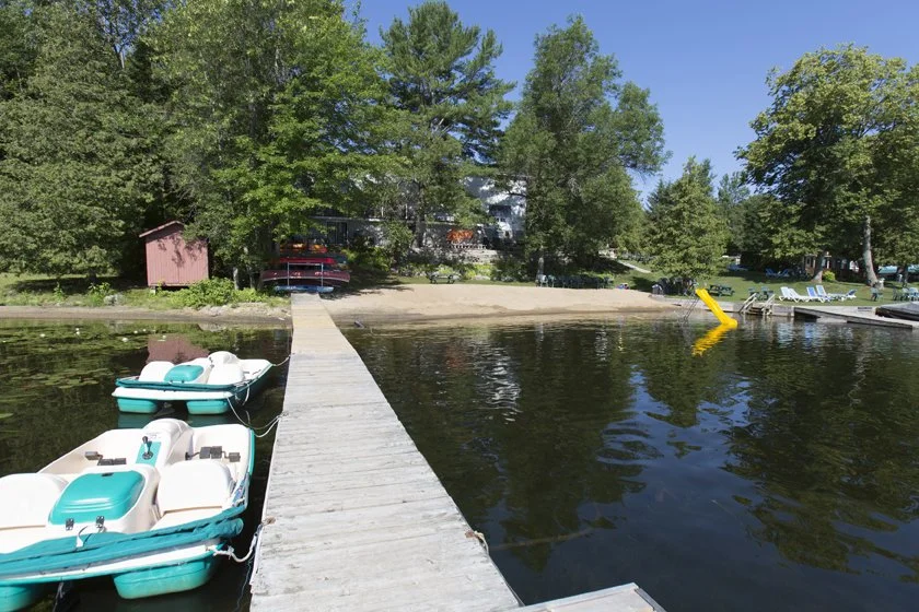 A dock extending into a lake with small pedal boats attached, leading to a sandy beach surrounded by trees and some lounge chairs on the grass.