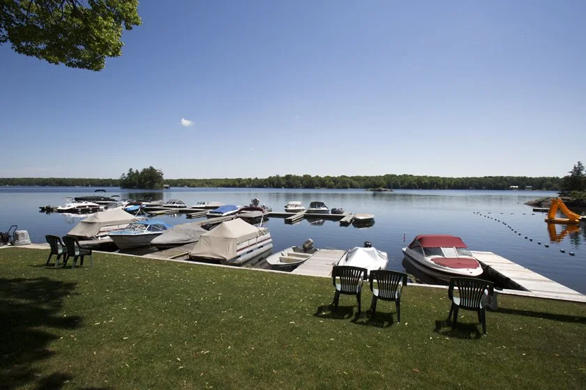 Boats docked along a lakeside with a grassy shore, outdoor chairs, and a clear blue sky.