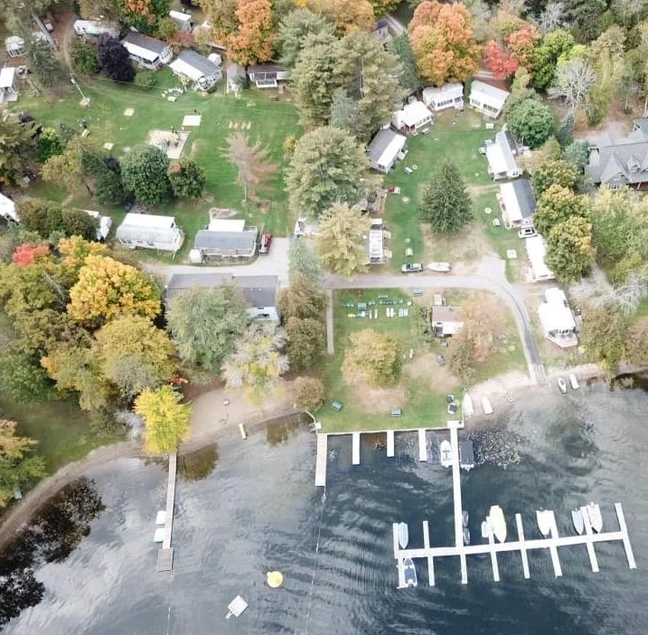 Aerial view of a waterfront area with houses, trees in fall colors, a small beach, and a boat marina docks on a lake.
