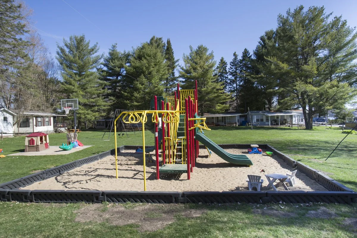 Playground with sandpit, swings, slide, and small plastic chairs in a grassy yard with trees and houses in the background.