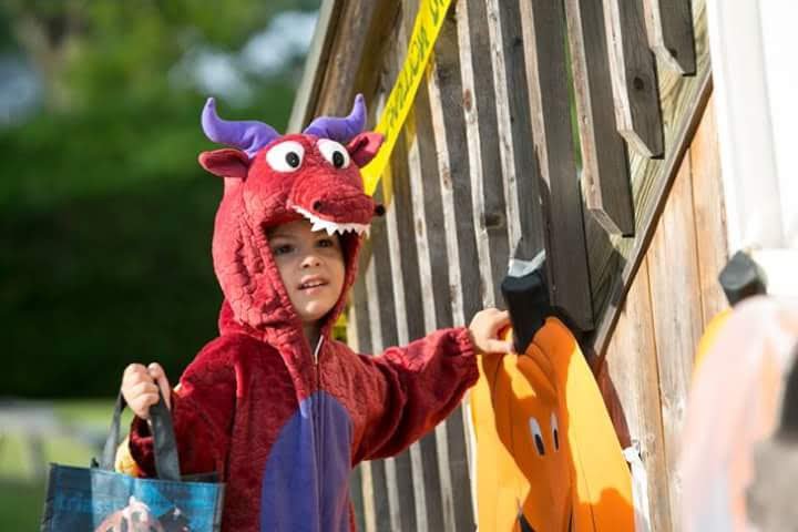 Young boy dressed in a dragon costume standing outside near a wooden fence with yellow caution tape. He is holding onto a black handle and looking to the side.