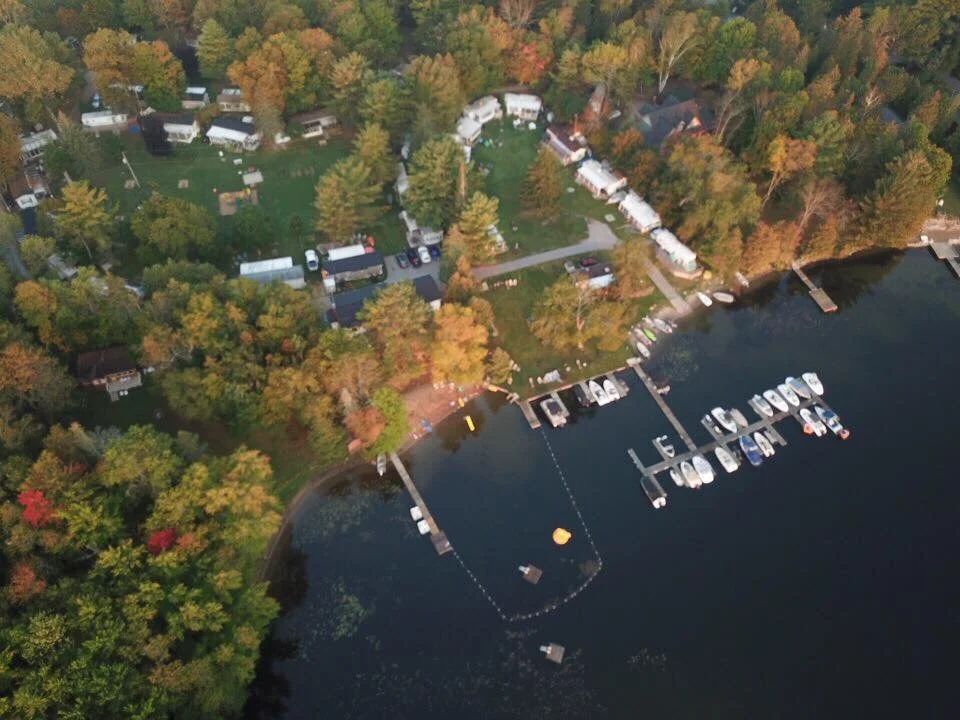 An aerial view of a marina with boats docked at a pier, adjacent to a wooded shoreline and scattered houses, with fall foliage on the trees.