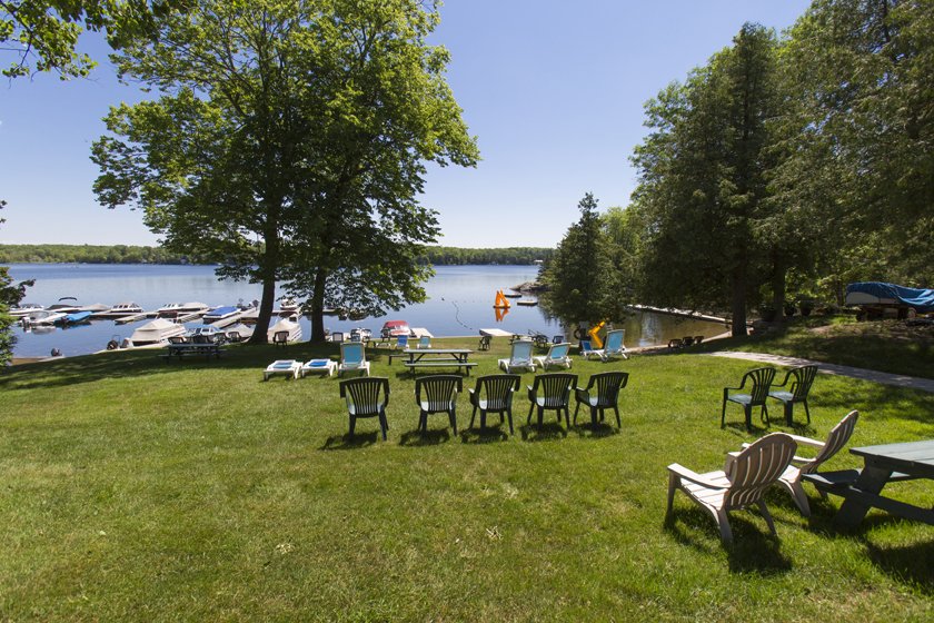 Lawn chairs and benches arranged on a grassy area by a lake with boats docked and trees in the background under a clear blue sky.