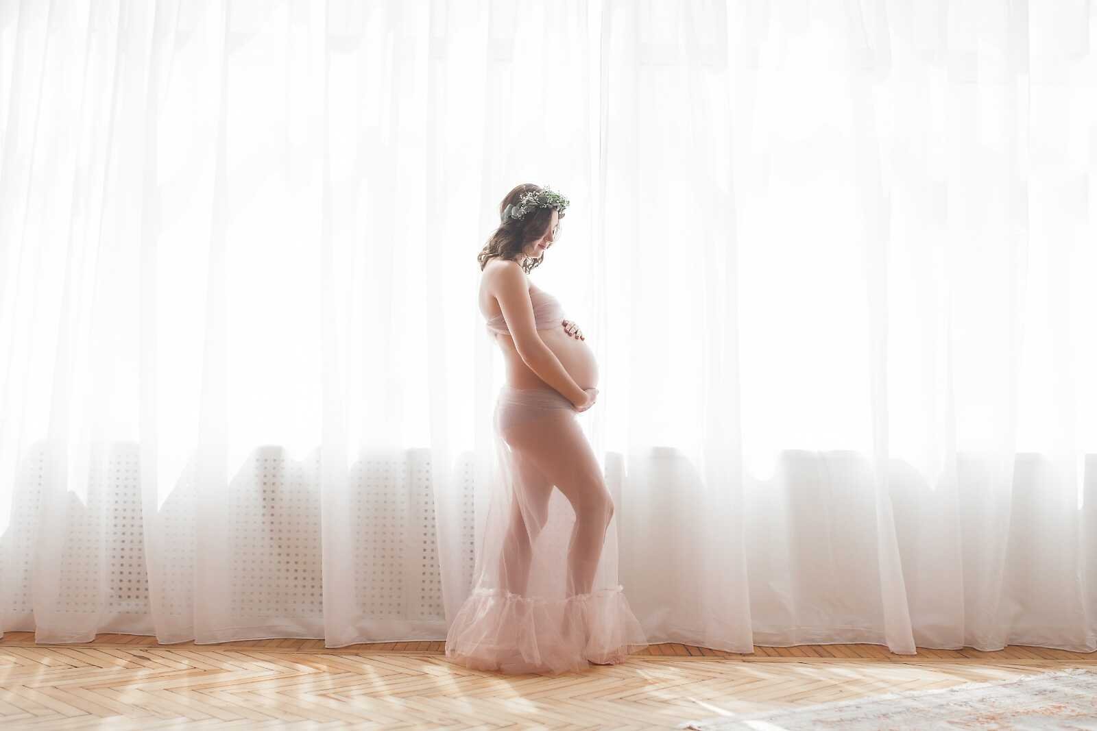 Nude maternity photos full body of pregnant woman in sheer fabric standing by bright window, soft natural light pregnancy portrait