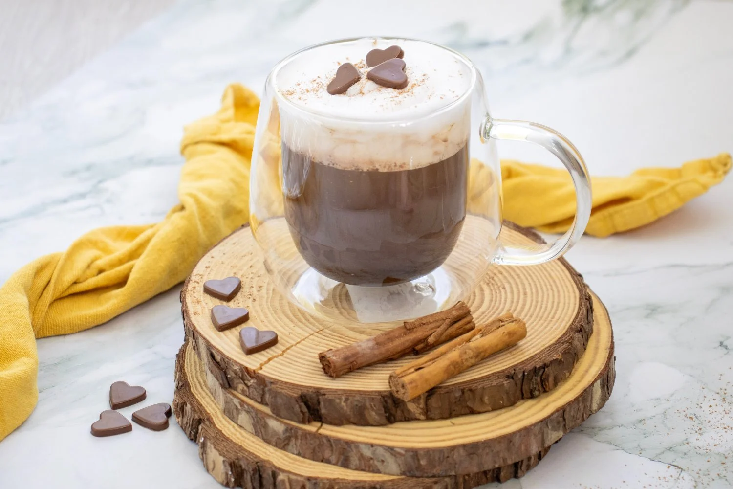 Pumpkin Spice Hot Chocolate topped with creamy foam, pumpkin pie spice, and chocolate hearts, served in a glass mug with cinnamon sticks.