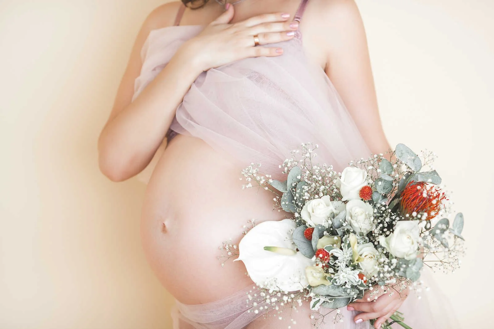 Nude maternity photos close-up of pregnant belly with sheer fabric and floral bouquet, soft neutral background pregnancy portrait