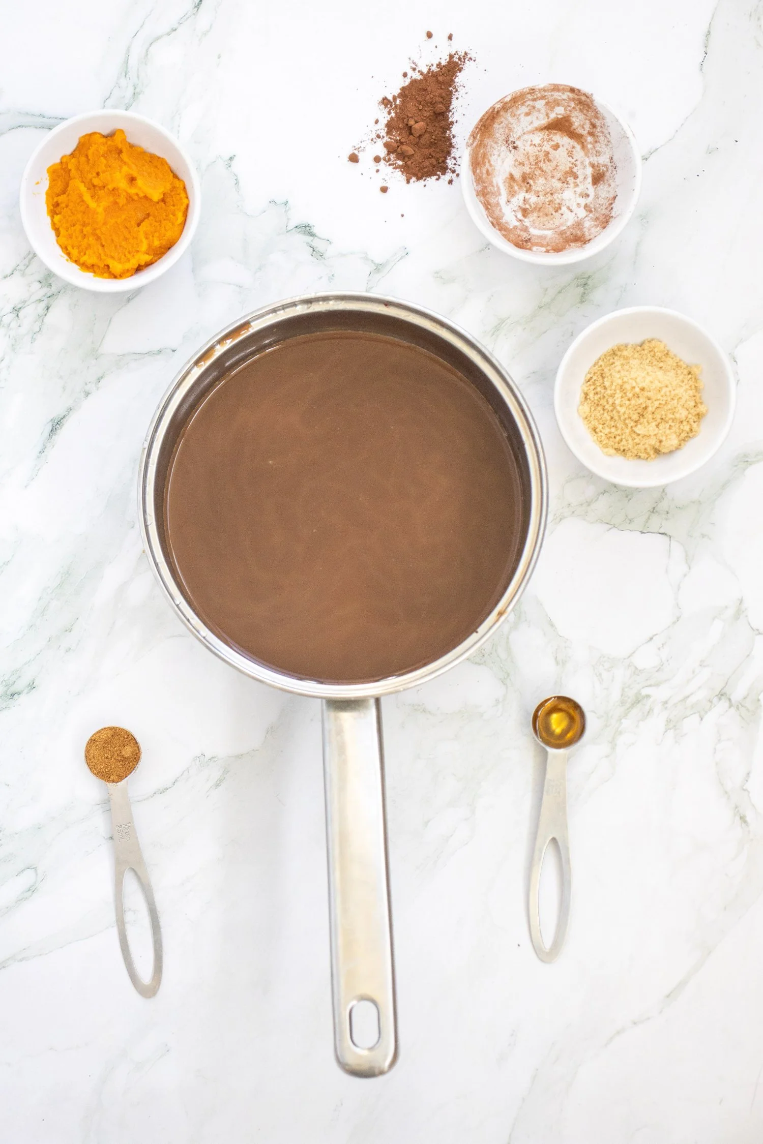 Pumpkin Spice Hot Chocolate mixture in a saucepan surrounded by pumpkin puree, cocoa powder, brown sugar, vanilla, and pumpkin pie spice.