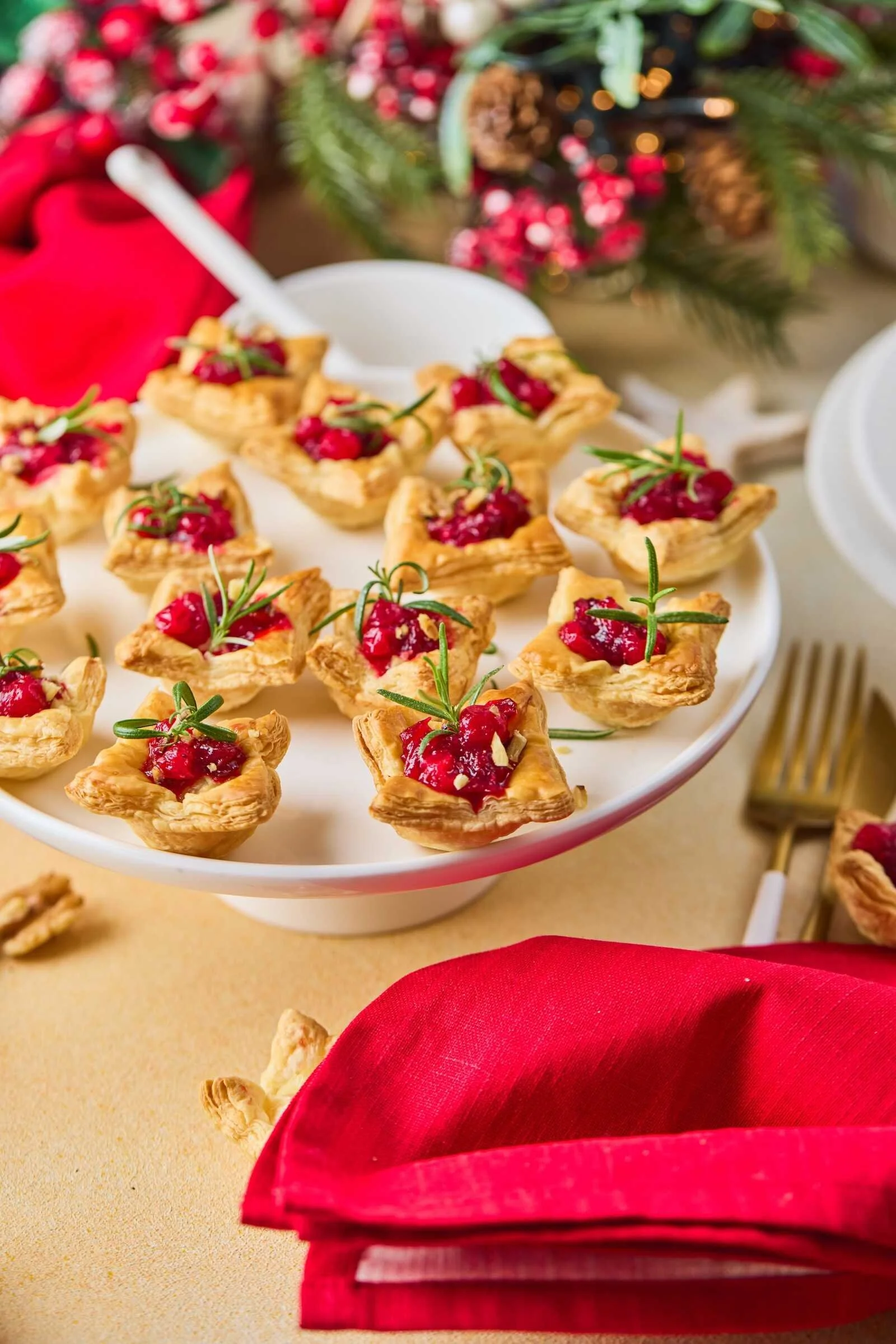 Holiday Heaven Bites served on a festive pedestal platter with flaky puff pastry, creamy Brie cheese, homemade cranberry topping, chopped walnuts, and fresh rosemary garnish.