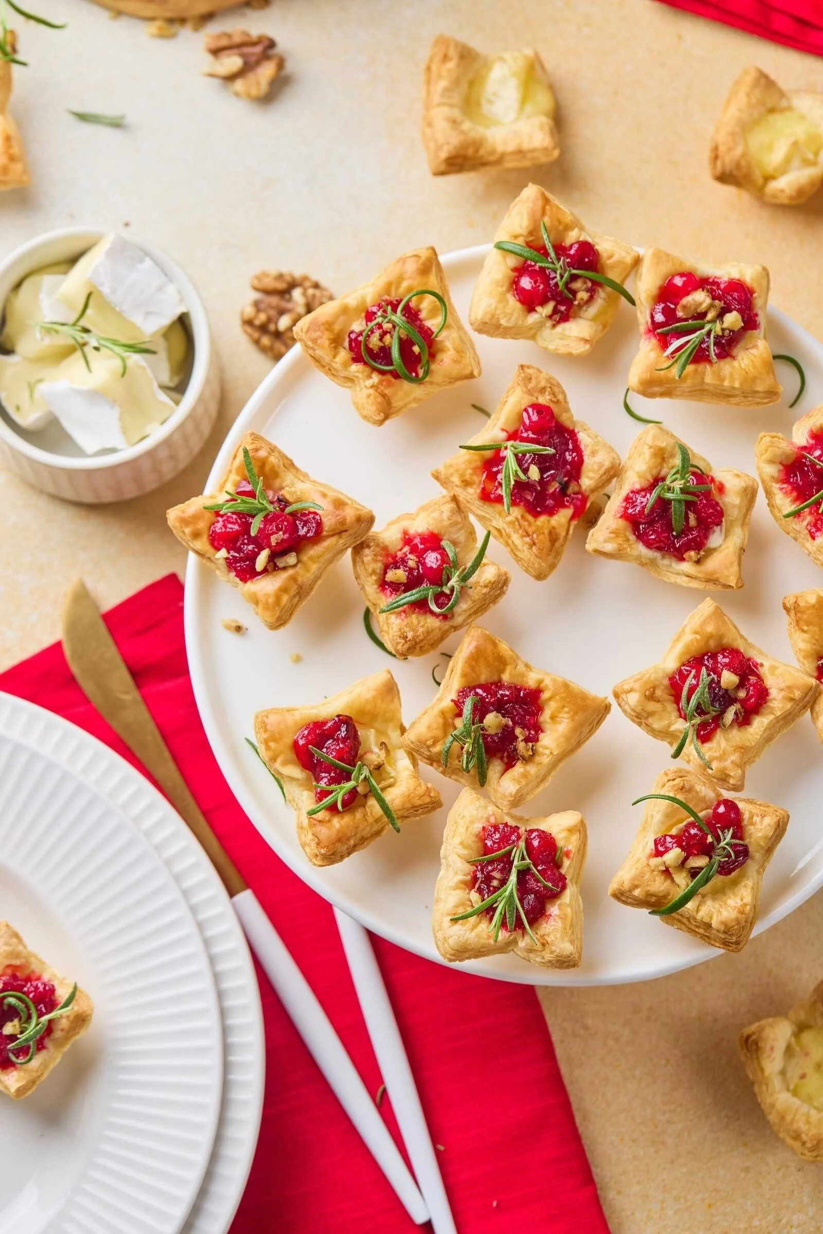 Overhead view of Holiday Heaven Bites with flaky puff pastry, creamy Brie cheese, homemade cranberry topping, chopped walnuts, and fresh rosemary served on a festive holiday platter.