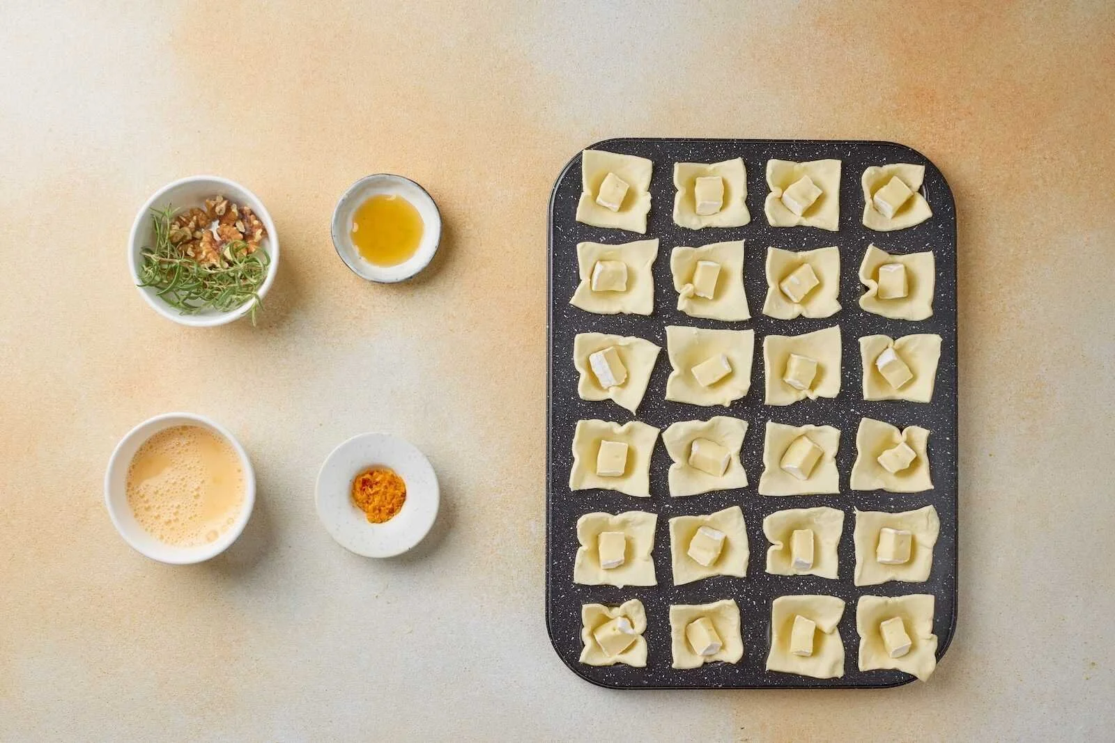 Holiday Heaven Bites prepared in a mini muffin pan with puff pastry cups, Brie cheese cubes, egg wash, honey, orange zest, rosemary, and walnuts before baking.