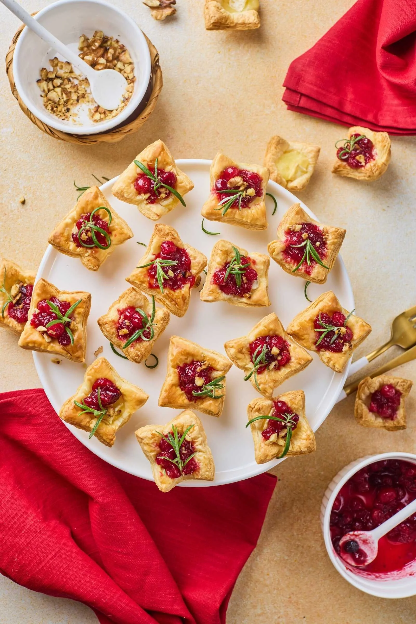Overhead view of Holiday Heaven Bites featuring flaky puff pastry, melted Brie cheese, cranberry topping, chopped walnuts, and fresh rosemary on a festive holiday serving platter.