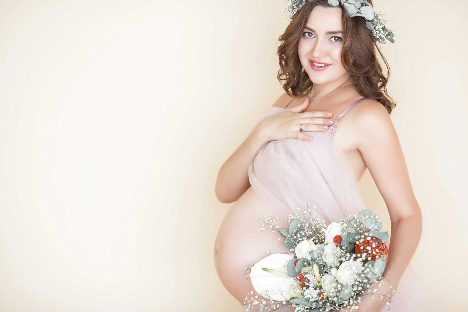 Nude maternity photos of pregnant woman in sheer fabric holding bouquet, soft neutral background pregnancy portrait