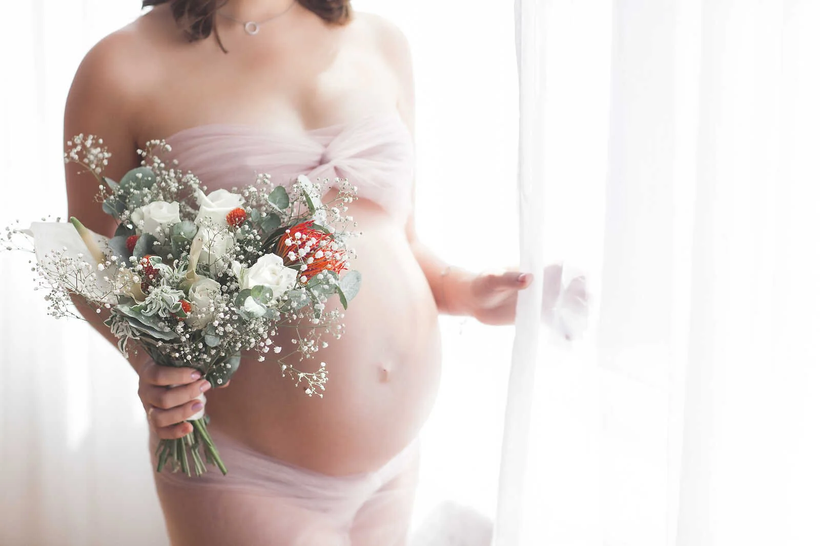 Nude maternity photos close-up of pregnant woman holding bouquet in soft natural light by window, artistic pregnancy portrait