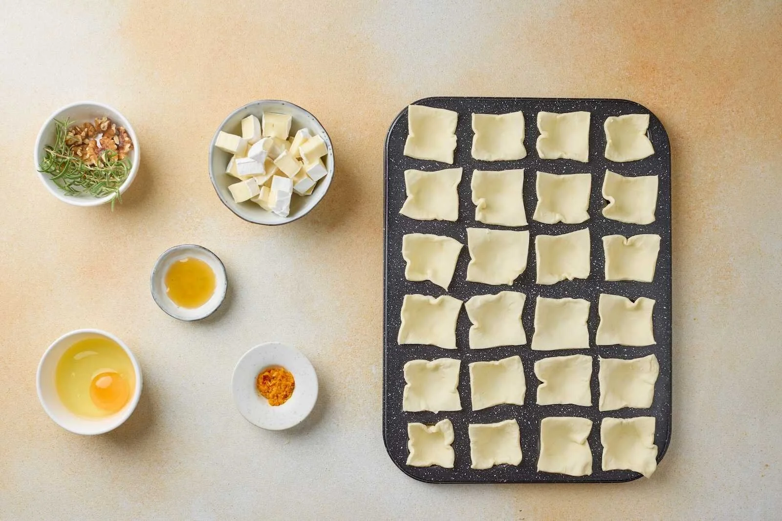 Puff pastry squares pressed into a mini muffin pan with Brie cheese, egg wash, rosemary, walnuts, honey, and orange zest prepared for Holiday Heaven Bites.