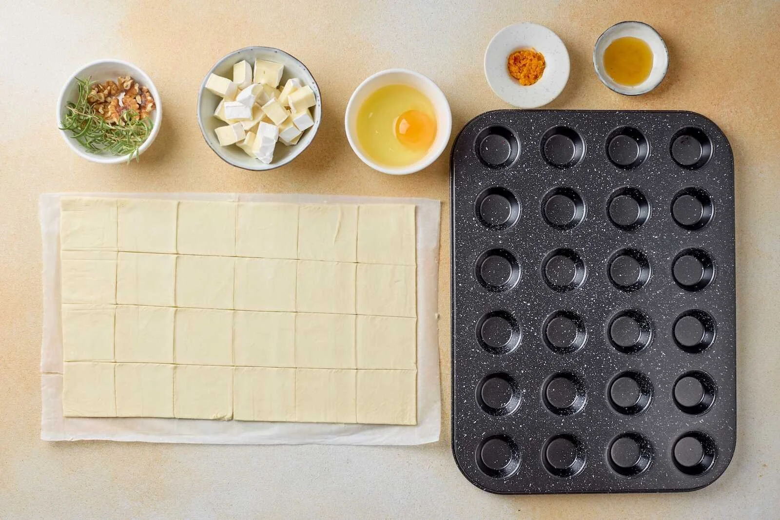 Puff pastry cut into squares alongside Brie cheese, egg wash, rosemary, walnuts, orange zest, honey, and a mini muffin pan for making Holiday Heaven Bites.
