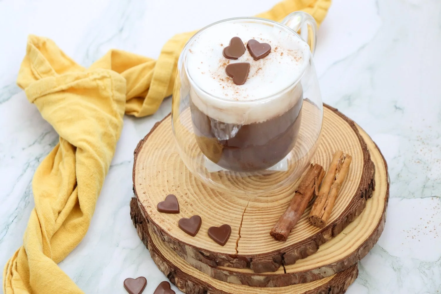 Pumpkin Spice Hot Chocolate topped with creamy foam, cinnamon, and chocolate hearts, served in a glass mug with cinnamon sticks.